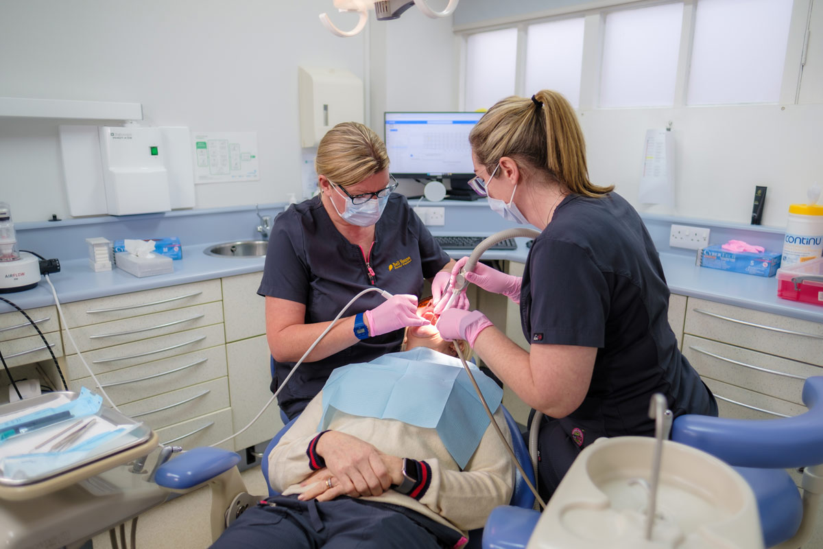 Two dental professionals wearing masks and gloves conduct a dental examination on a patient in a brightly lit, modern dental clinic, surrounded by dental equipment and monitors.