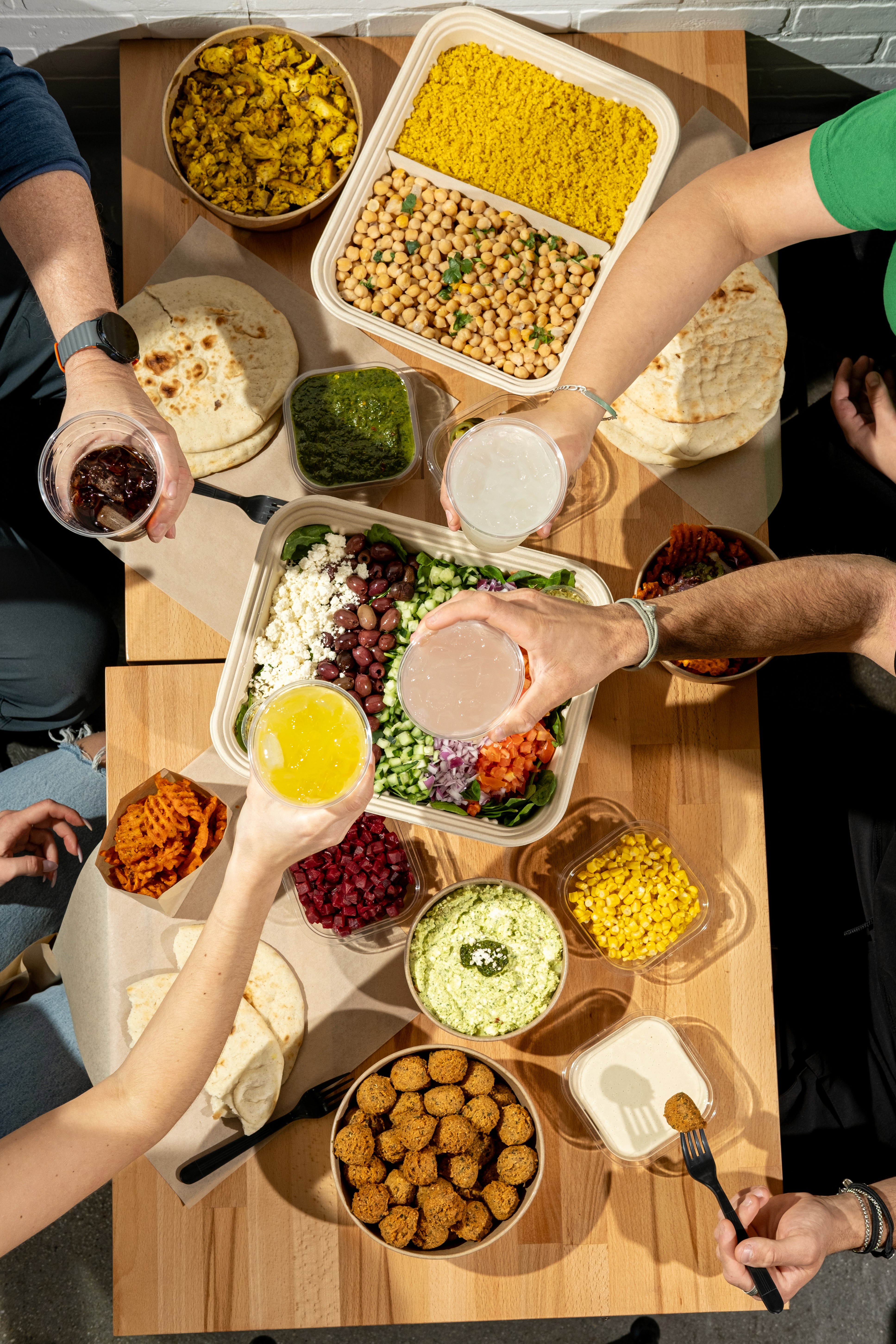 top down shot of table with various food in containers and 4 hands cheersing with drinks