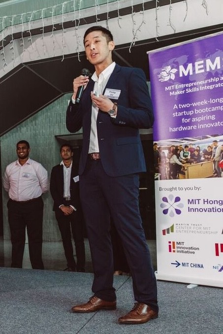 Man in a dark suit speaking into a microphone on stage at an event, with a banner in the background promoting MIT Entrepreneurship and Maker Skills Integration. Other individuals are standing nearby, and the event appears to be related to a startup bootcamp for aspiring hardware innovators.