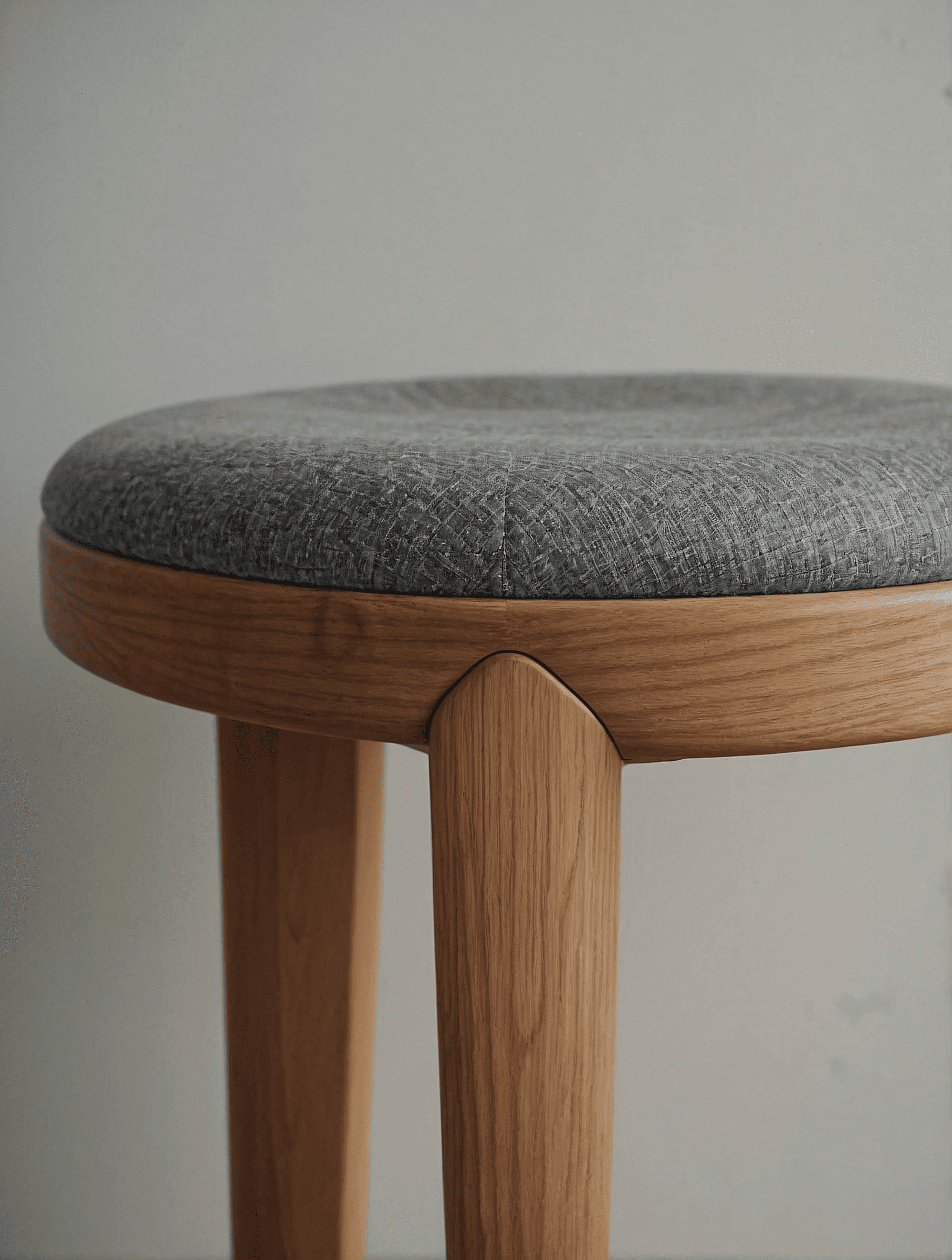 Minimalist wooden stool with a gray cushioned seat and light oak legs, showcasing clean lines and precise joinery in a neutral setting.
