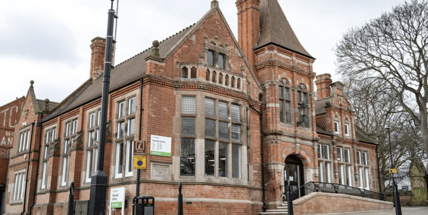 A photo of the front of Hucknall Library. It is a Victorian building designed in a renaissance revival style. It is red brick, with detailed windows and a sign outside.