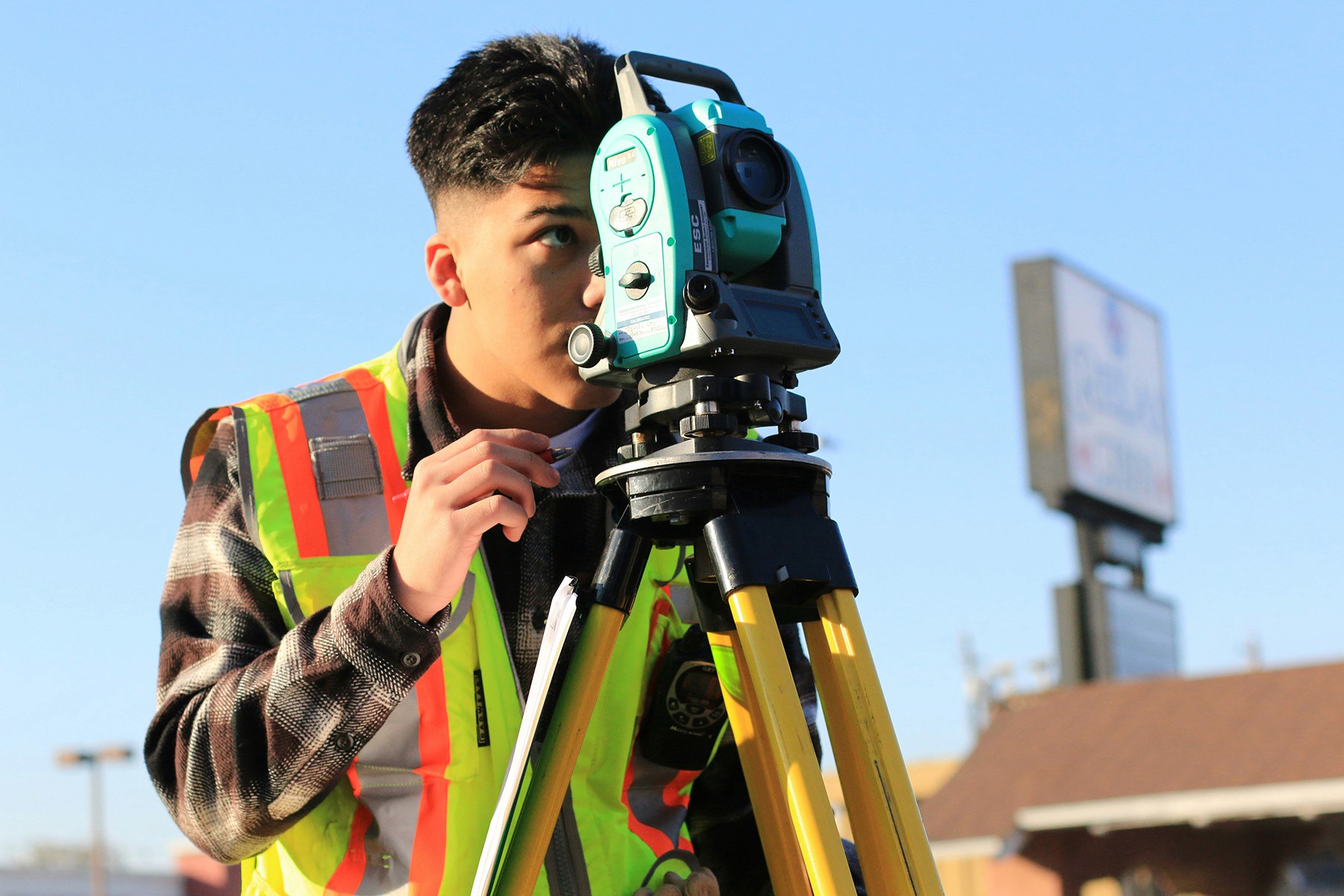 A young land surveyor, wearing a safety vest, looks through surveying equipment on a tripod.