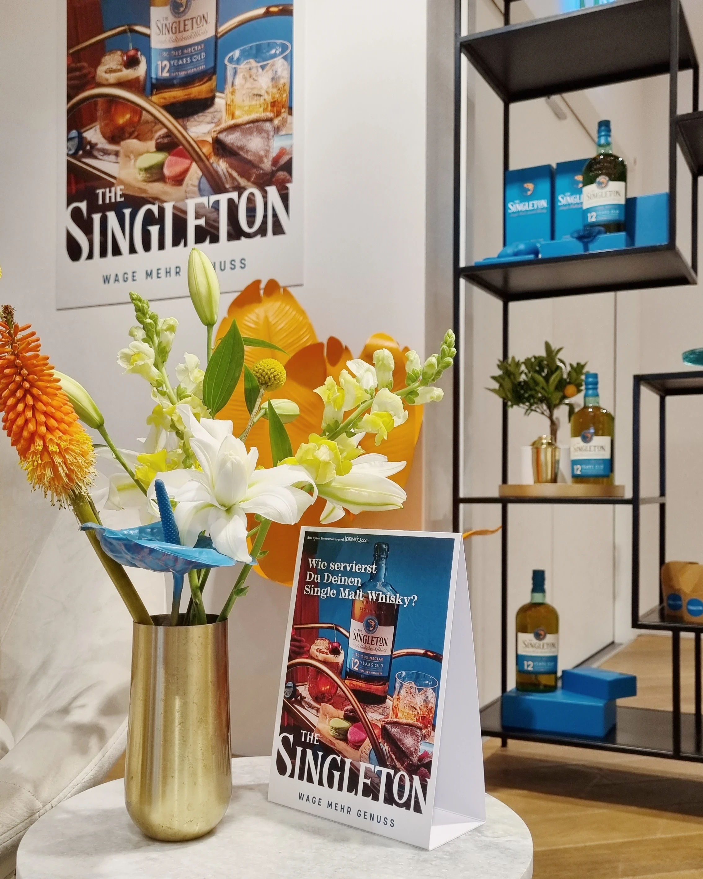 Gold vase with flowers and Singleton table display in front of the campaign poster and shelf.