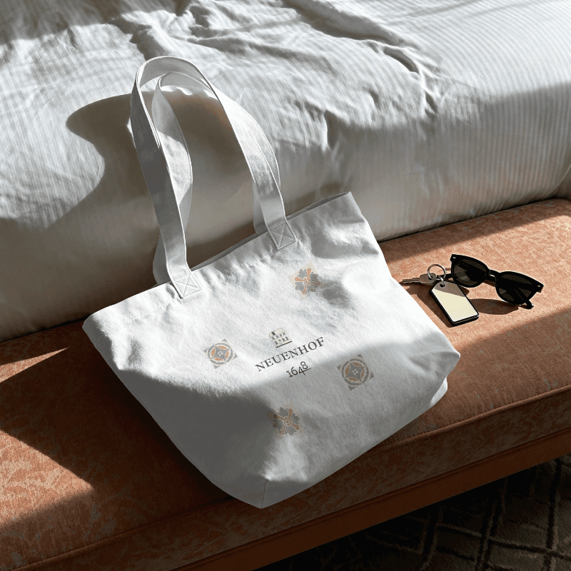 Weiße Stofftasche mit Logo und ornamentalen Elementen  am Fussende eines weiß bezogenen Bettes, liegend auf einer Bank mit organemem Stoff