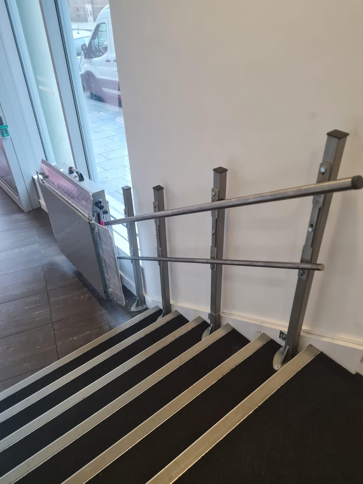View from upper landing looking down straight staircase with incline platform stairlift folded at top — stainless steel balustrade and anti-slip treads in commercial building