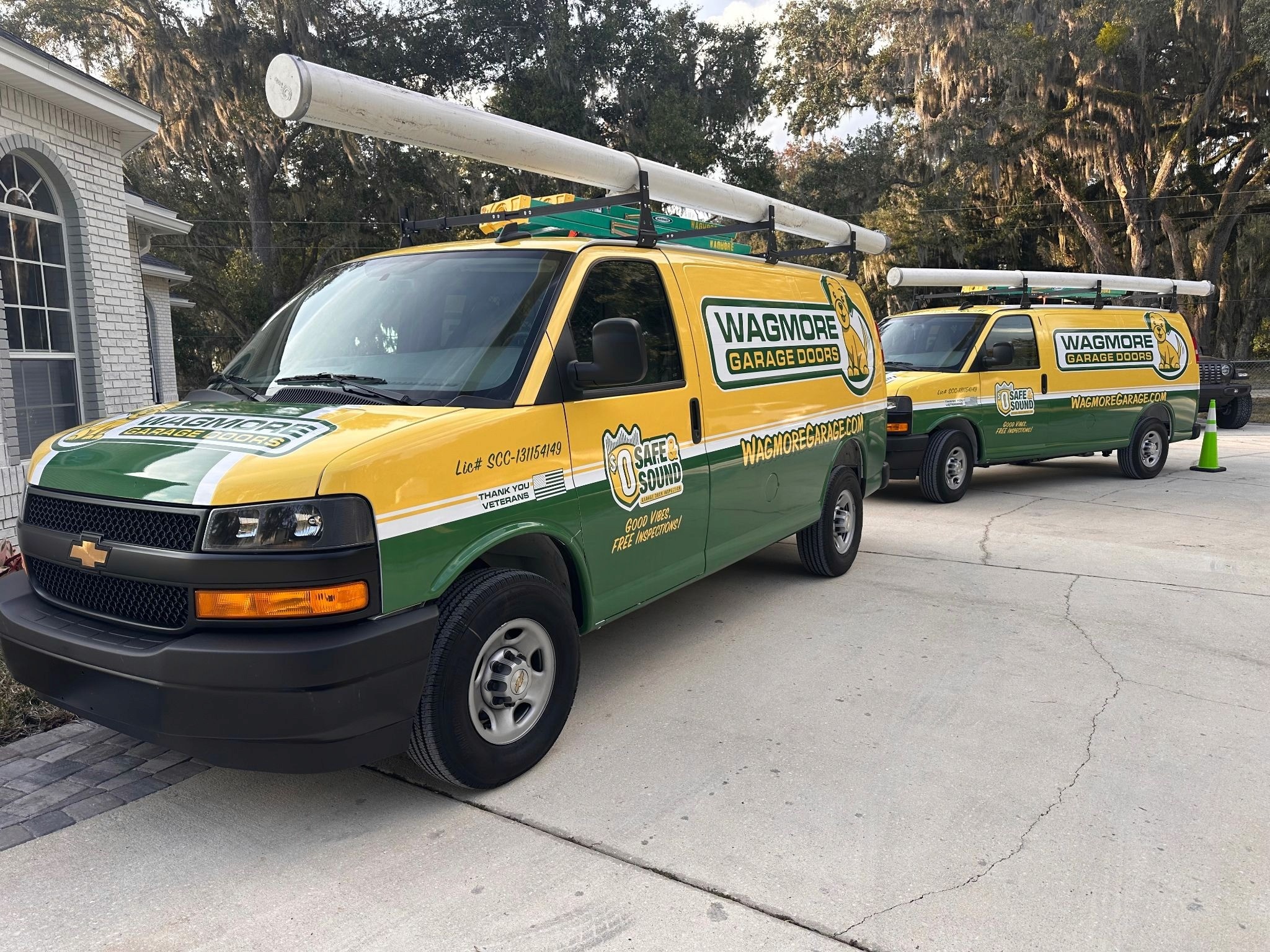 Two yellow and green vans with "Wagmore Garage Doors" branding are parked on a driveway. Both vehicles have ladders on top, conveying a sense of service.