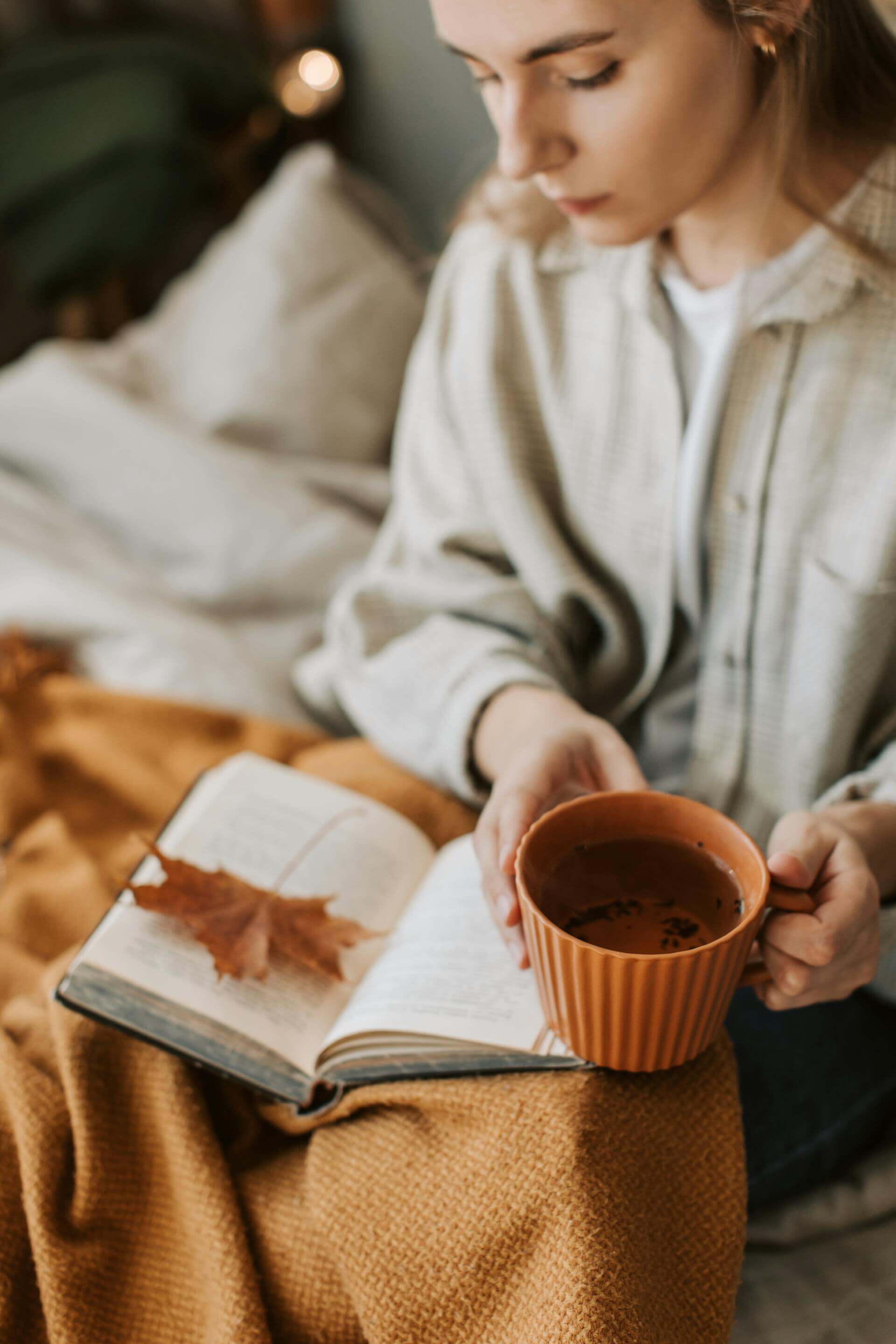 A woman sits on a bed, engrossed in a book while sipping tea, creating a cozy and serene atmosphere