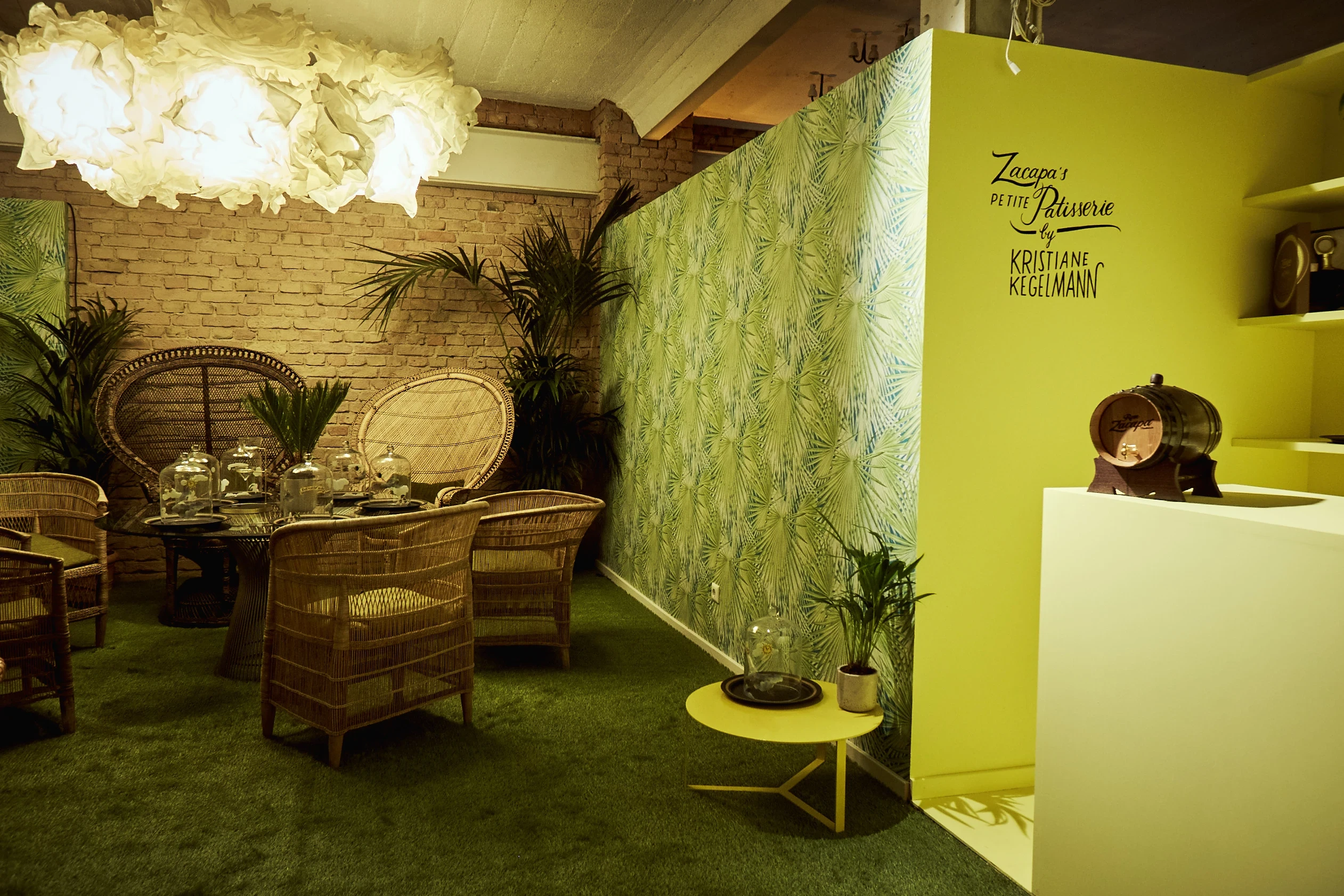 Zacapa lounge at DIAGEO BCB17 with rattan peacock chairs on green carpet, palm-print wallpaper, a paper cloud lamp and a yellow display unit.