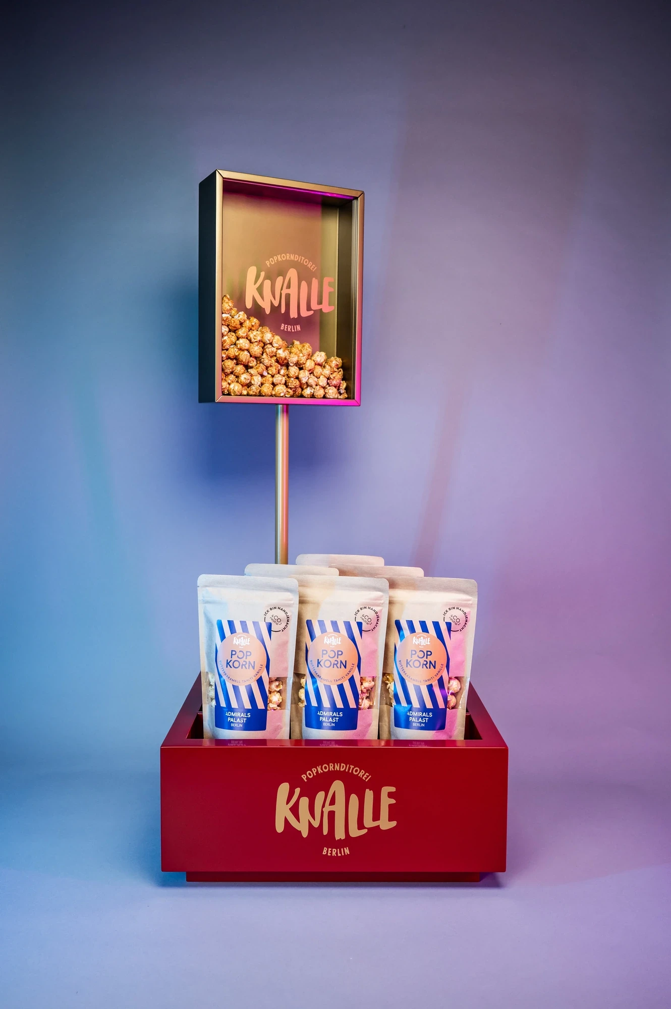 Knalle Popcorn POS display prototype with a red base, three branded popcorn pouches and a gold-framed acrylic logo box raised on a slender stem.