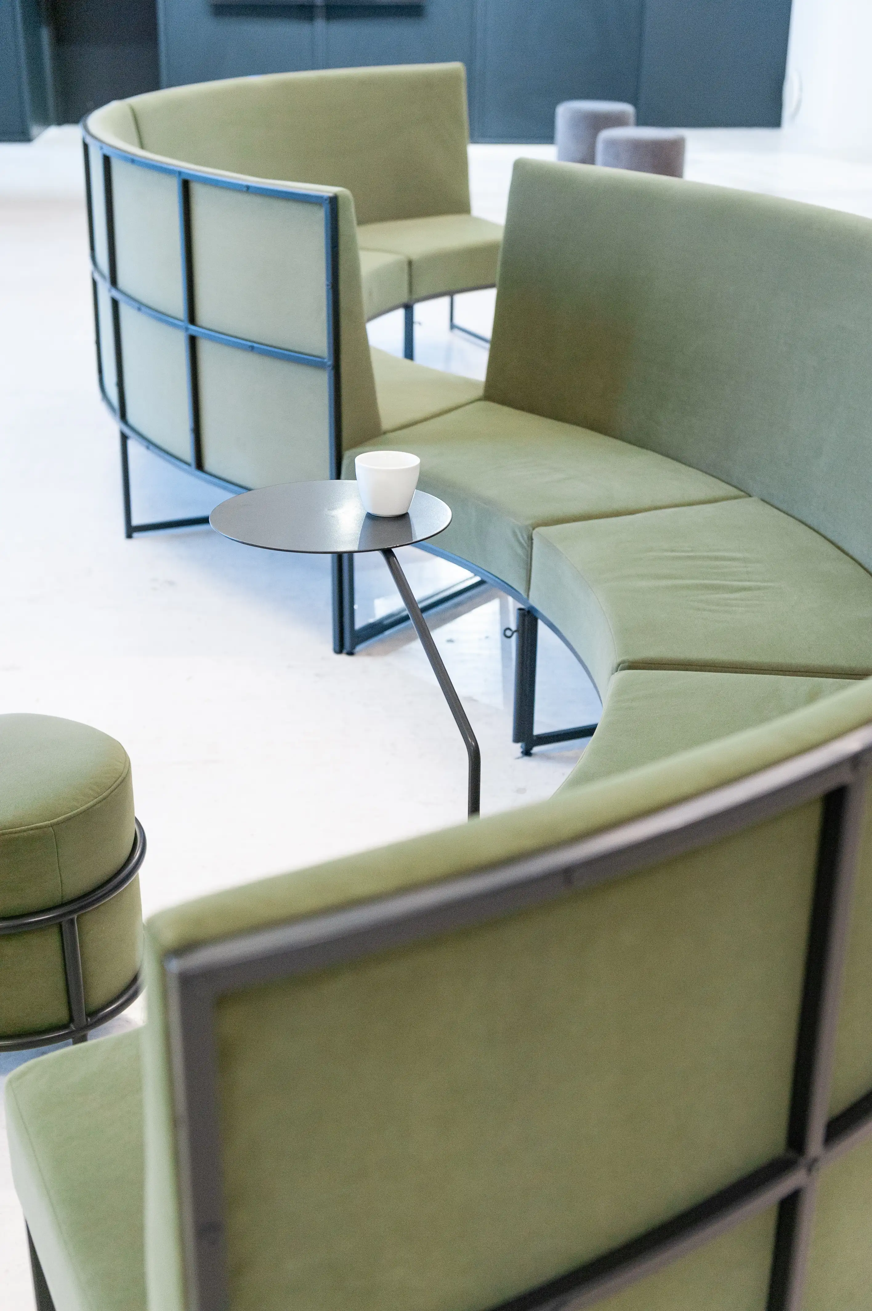 Detail of curved sage-green velvet benches in black steel frames at LA RED with a small round black side table holding a white coffee cup.