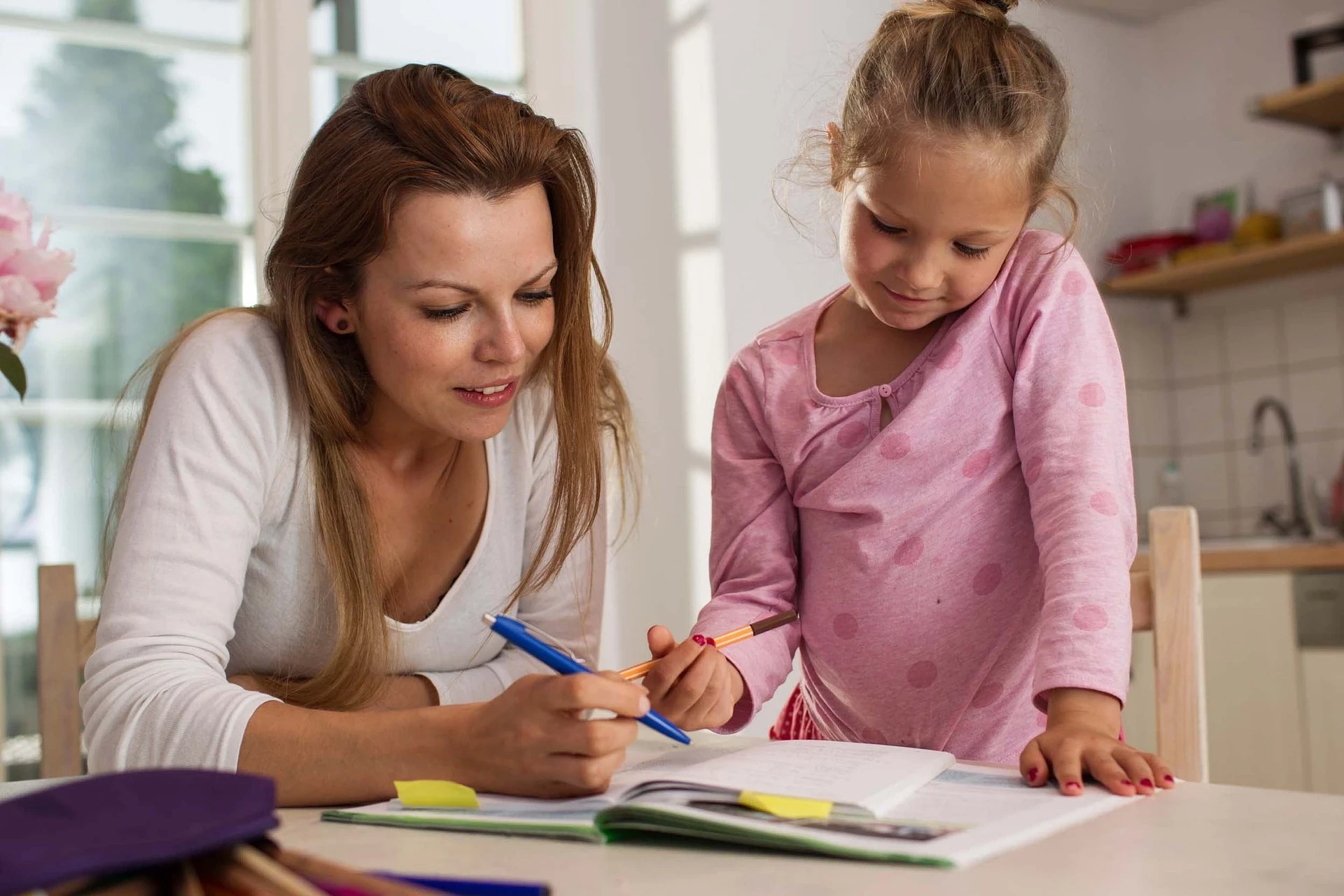 Mulher adulta e menina pequena trabalhando juntas em uma mesa, onde há cadernos e canetas. A mulher, de cabelos castanhos e blusa branca, está sorrindo e apontando para algo no caderno com uma caneta. A menina, com blusa rosa e cabelo preso, observa atentamente. A cena transmite um momento de aprendizado e cuidado.