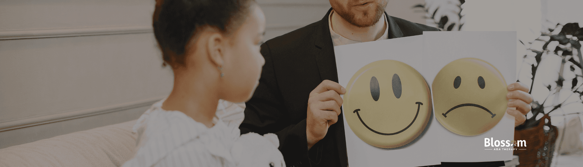 Therapist holding up a card with happy and sad faces while a girl with a teddy bear looks at it