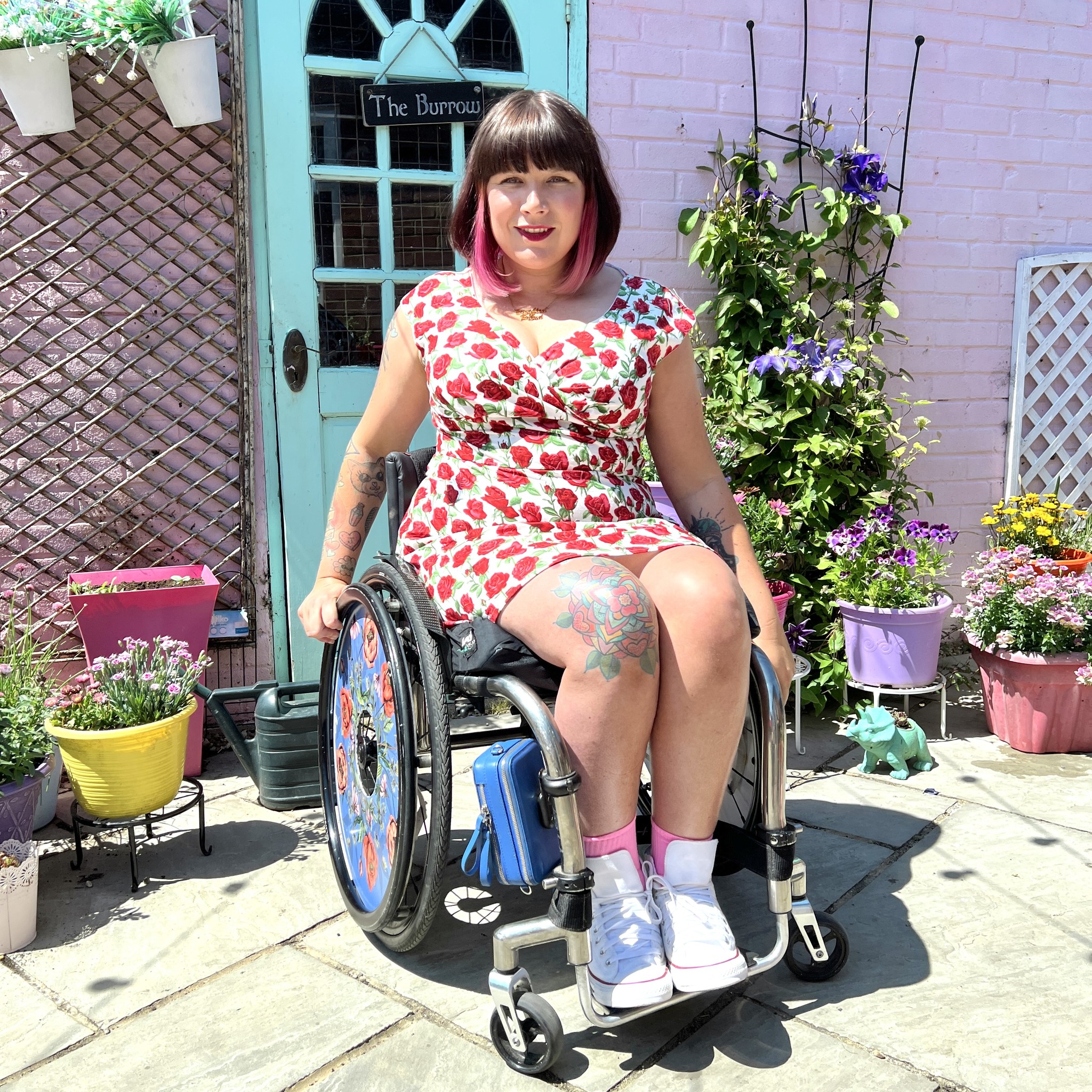 Nina, a wheelchair user with black and pink hair, is posing in front of her front door wearing a dress covered in red roses, and white converse. The door is mint against a pink wall and there are lots of potted plants all around.