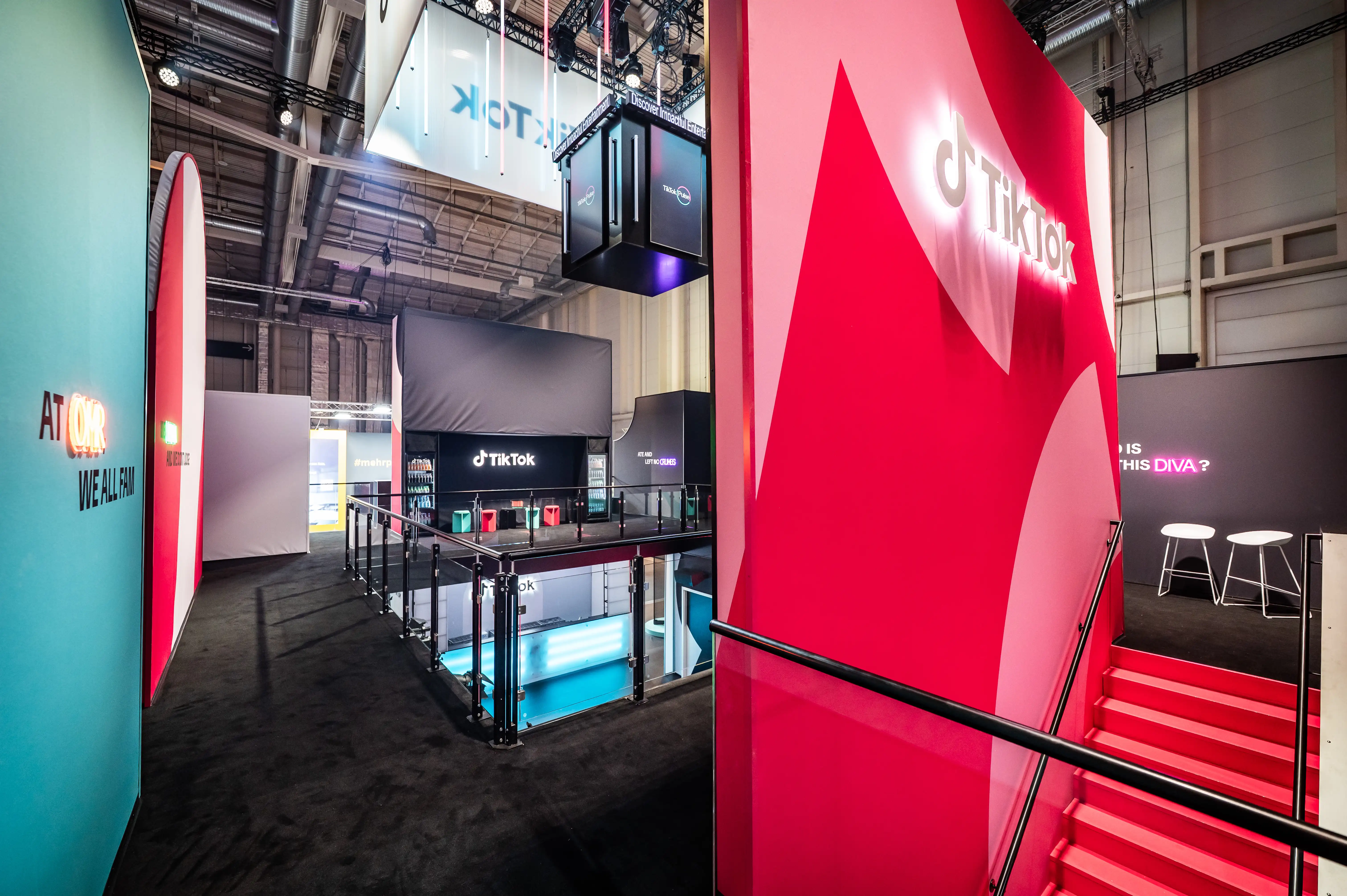 Upper deck of the TikTok booth with glowing logo wall, red staircase and turquoise panels around the gallery.