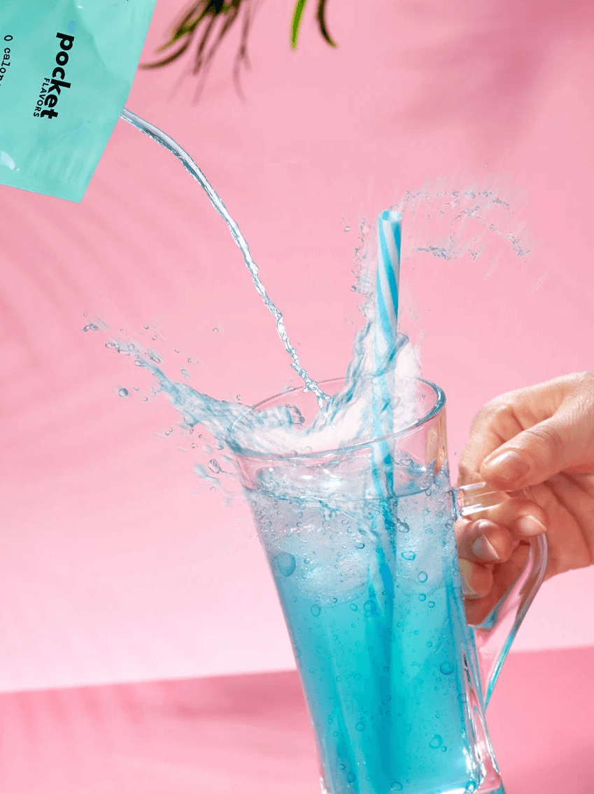 A hand pours blue liquid from a pocket-shaped container into a tall, clear glass with a striped straw, causing vibrant splashes against a light pink background, showcasing a lively motion and animation scene from a commercial.