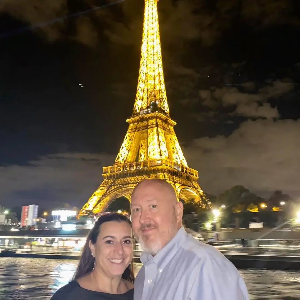 A couple stands close together in front of a brightly illuminated Eiffel Tower at night, capturing a romantic atmosphere reminiscent of a dream travel experiencefrom Your Bucket List Company, Florida.