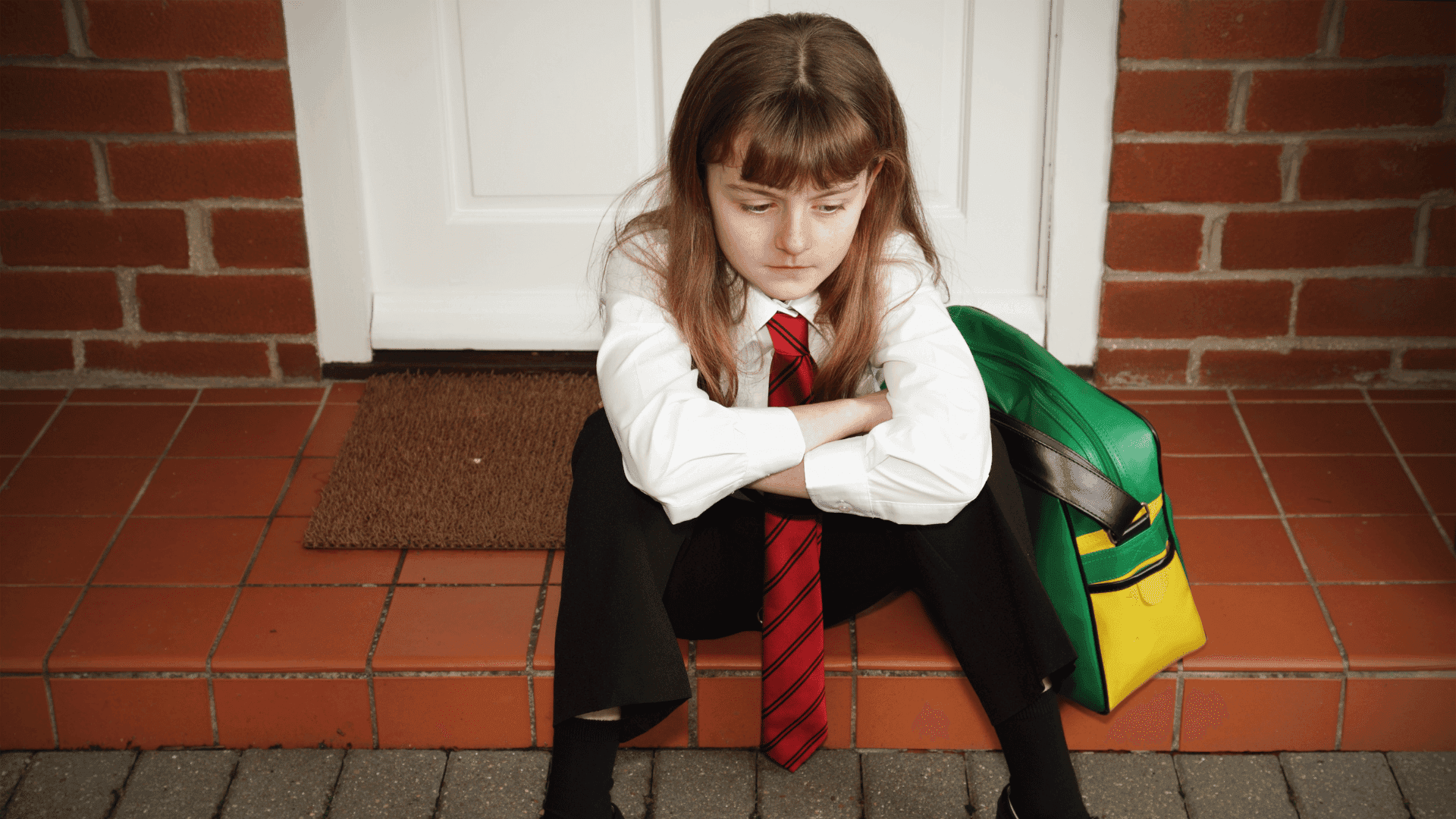 A girl sitting on her front door step, looking sad after a day at school.