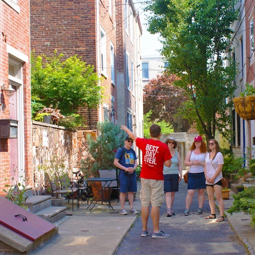 A group of people stand in an alleyway with a tour guide in a red shirt pointing at something. Brick buildings and greenery surround them.