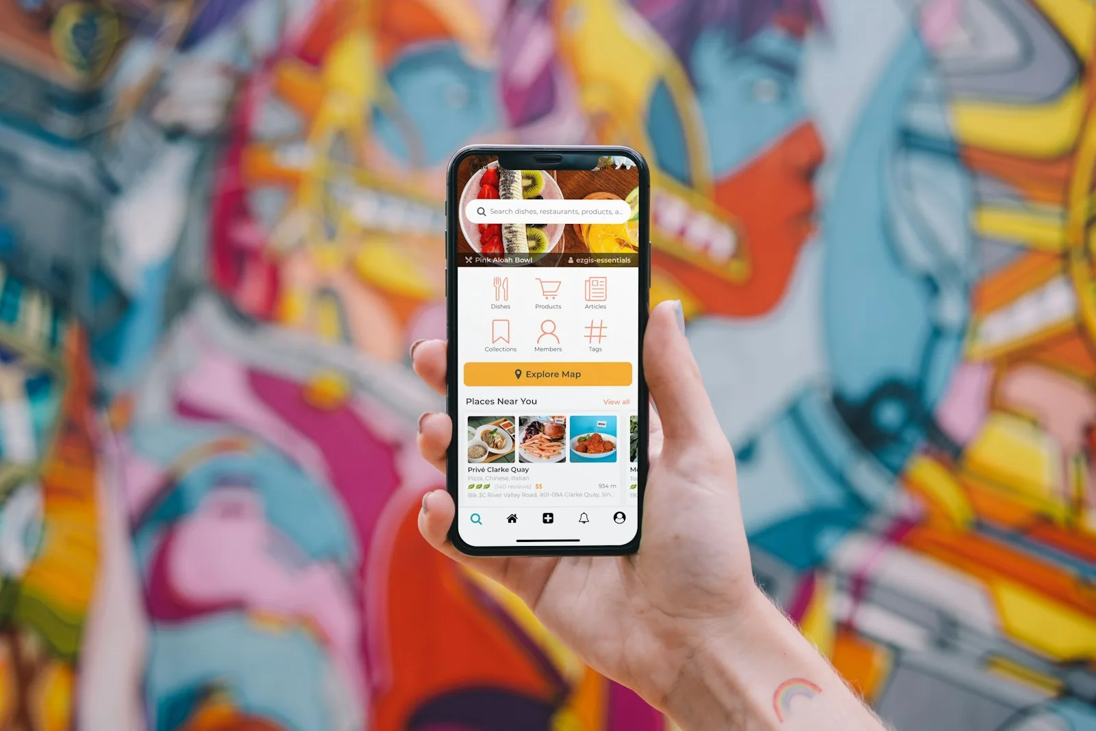 Hand holding a smartphone showing a food discovery app with search, categories and nearby places, in front of a colorful street mural