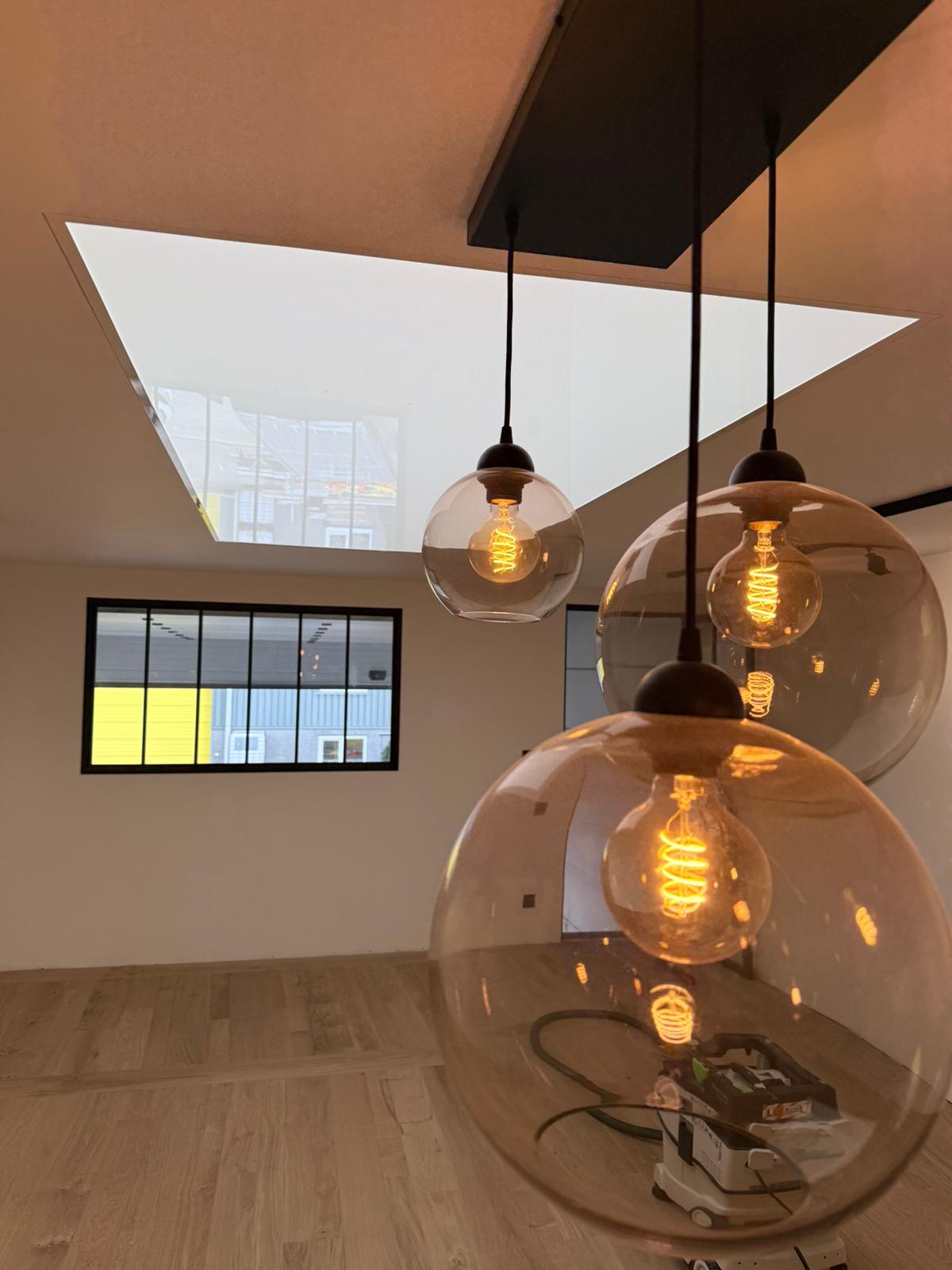 Grote bolvormige hanglampen met zichtbare Edison-achtige filamenten in een dubbelhoge ruimte, met op de achtergrond een raam met donker frame en subtiele reflecties van geel en blauw licht, wat kleur toevoegt aan de warme verlichting.