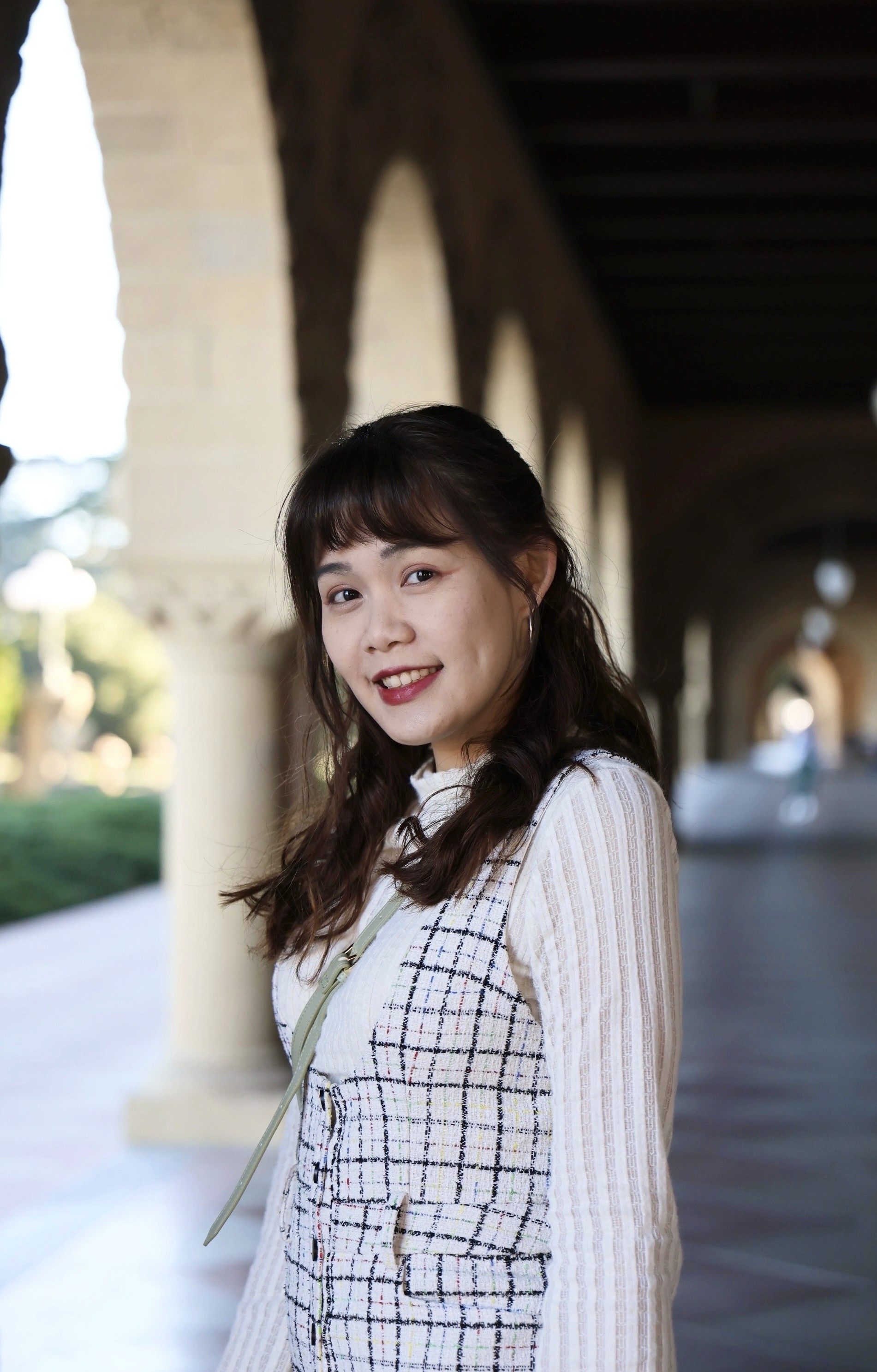 Headshot of a young woman with shoulder-length brown hair, wearing a white sweater and a checkered pinafore dress. She's smiling and looking at the camera.