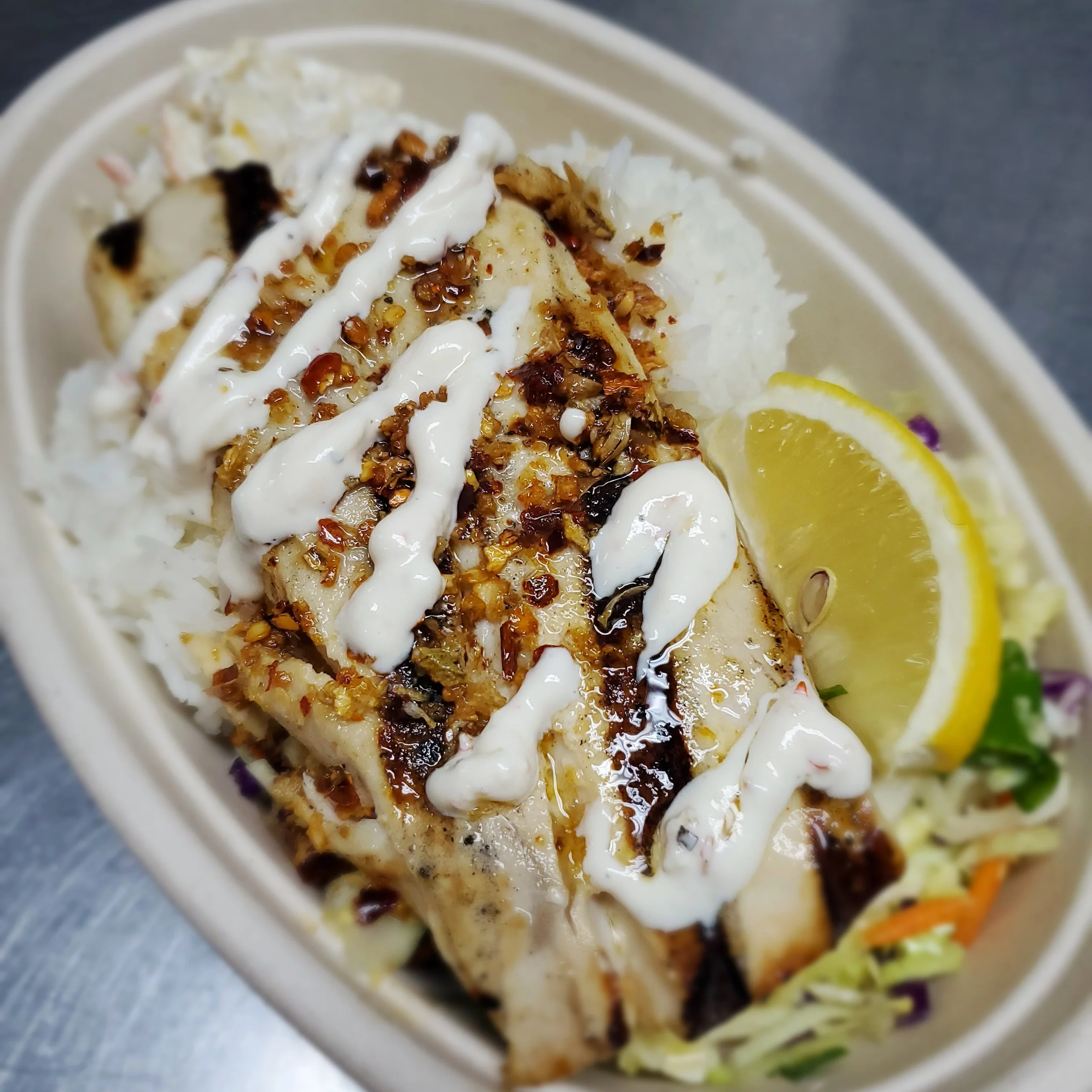 A char-grilled Mahi Mahi filet topped with garlic cream sauce and chili flakes, served in a bowl with two scoops of rice, macaroni salad, and Asian slaw with a lemon wedge.