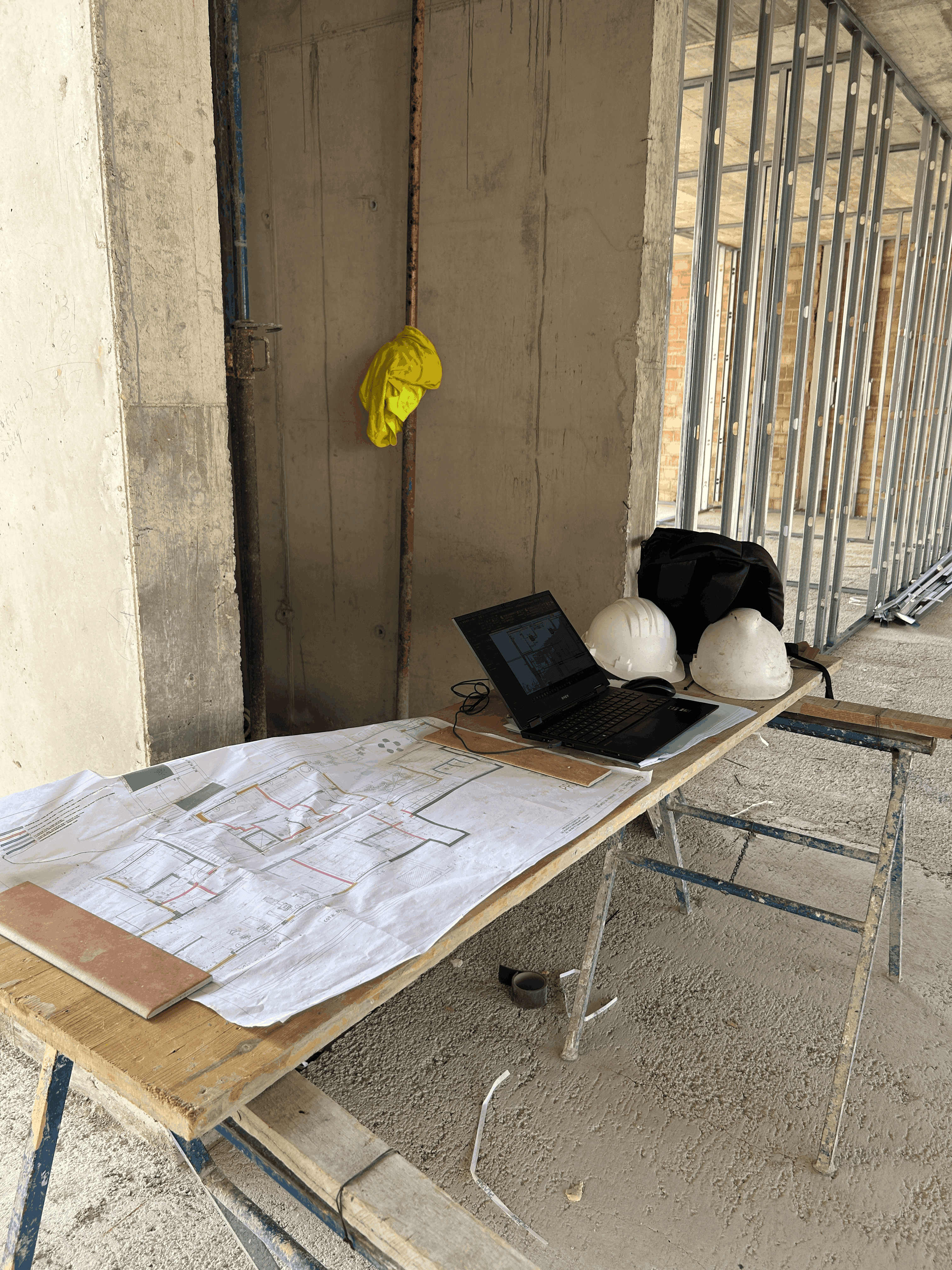 Puesto de trabajo de dirección de obra en un edificio en construcción, con planos técnicos, ordenador portátil y cascos de seguridad sobre una mesa provisional.
