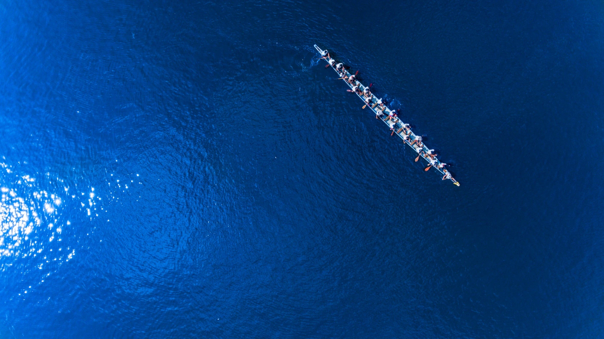 Aerial view of a long rowboat with multiple people rowing across the blue water.