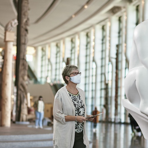 一位戴着口罩的女士在一个宽敞、现代的画廊里观赏一个大型雕塑，画廊天花板很高，窗户很大。