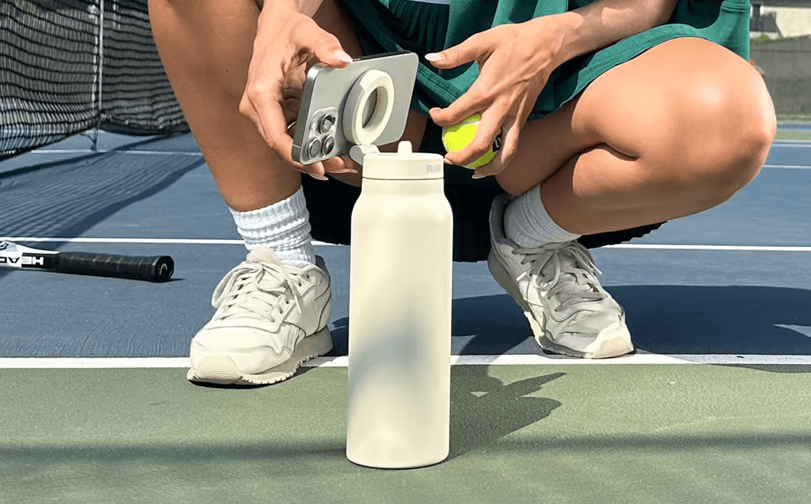someone on a tennis court squatting down to attach their phone to their ringo waterbootle only showing their knees and feet in the frame of the image.
