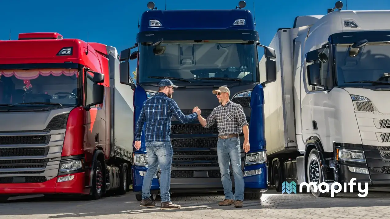 Two men in casual plaid shirts and hats exchange a fist bump in front of a red, a blue, and a white semi-truck, conveying a sense of teamwork and camaraderie.