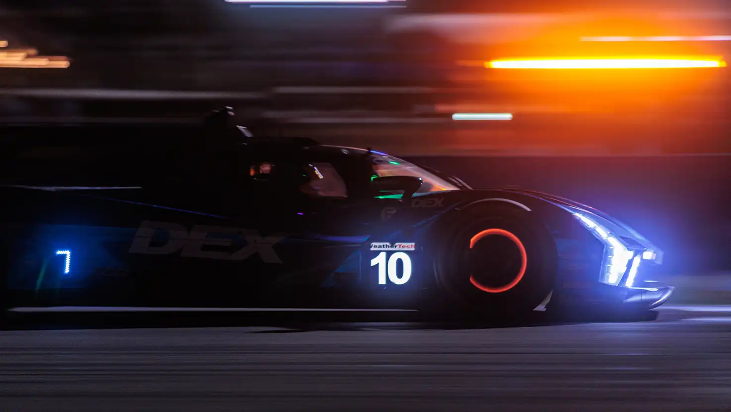 A race car speeds down a dimly lit track at night, with bright headlights illuminating the path ahead.