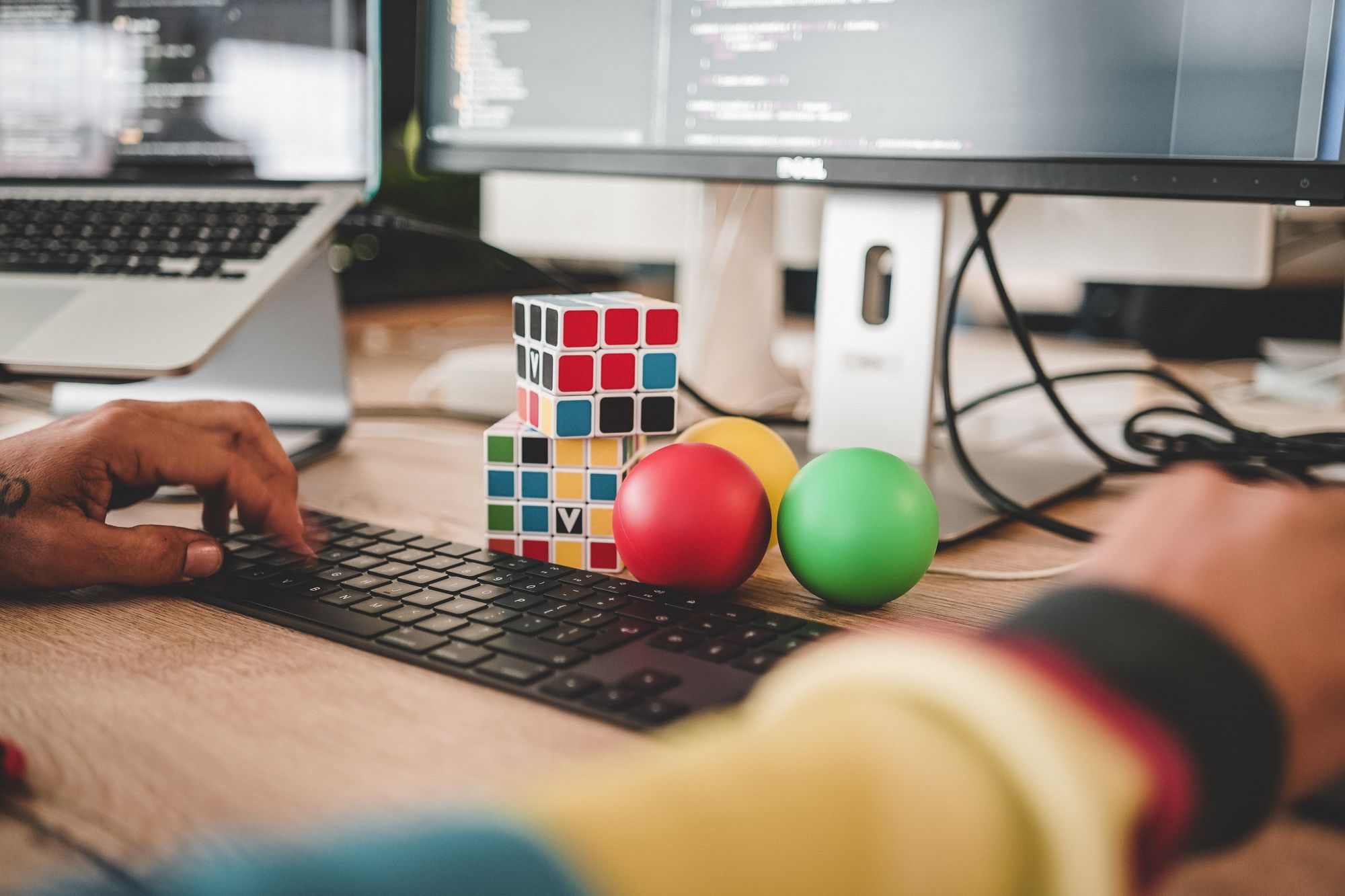 Two colorful Rubik's cubes beside stress balls above a keyboard while person is working