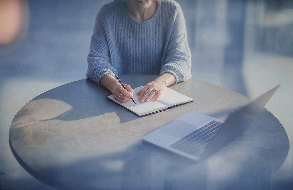 Profissional escrevendo em caderno ao lado do notebook em mesa de escritório, com reflexo de vidro em primeiro plano. Blog post Inspira.