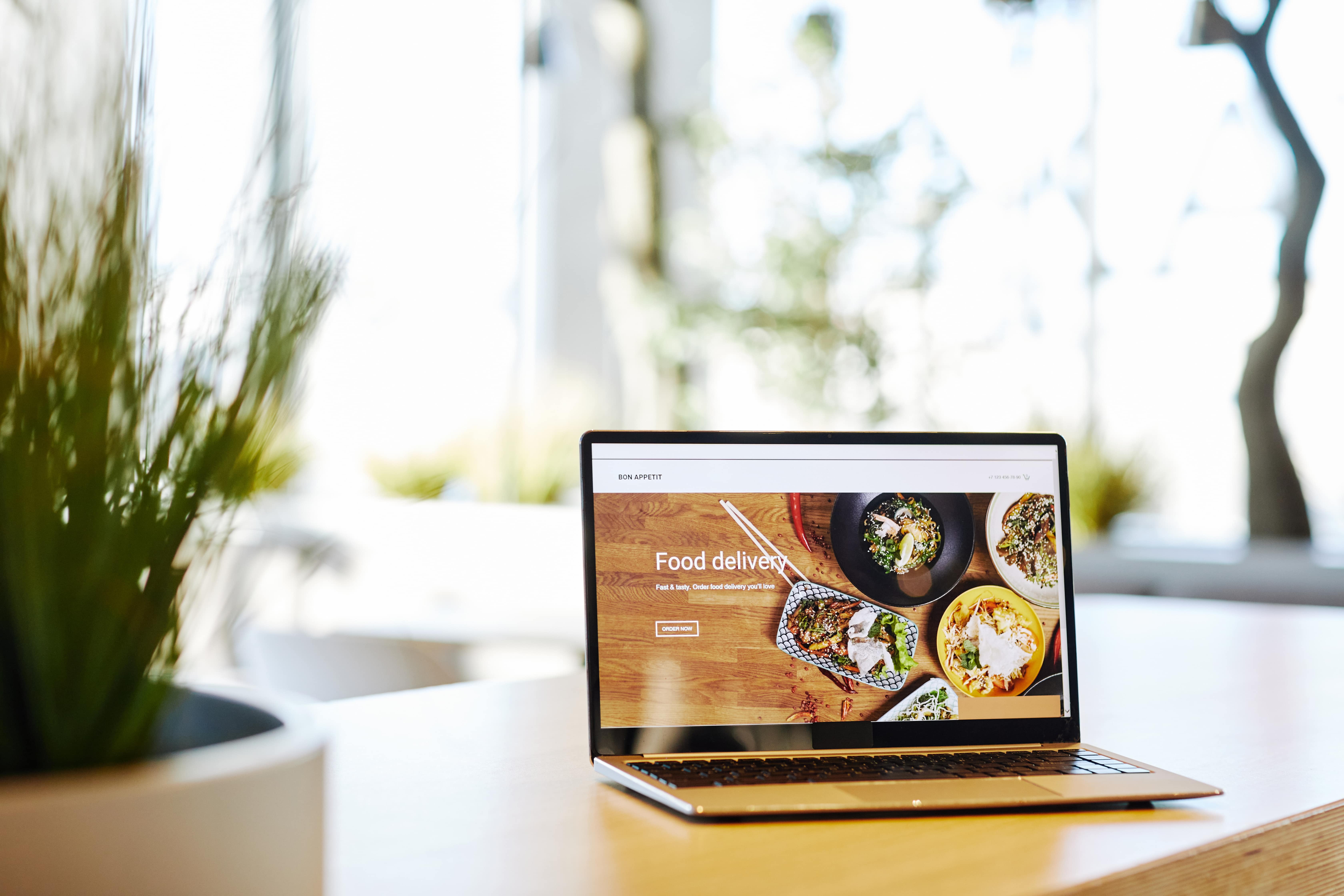 An open laptop sits on a wooden desk in a modern, brightly lit office space, with a blurred green plant in the foreground. The laptop screen displays a user-friendly website for a food delivery service with the headline "Food delivery," an "ORDER NOW" button, and high-quality images of various appetizing meals.