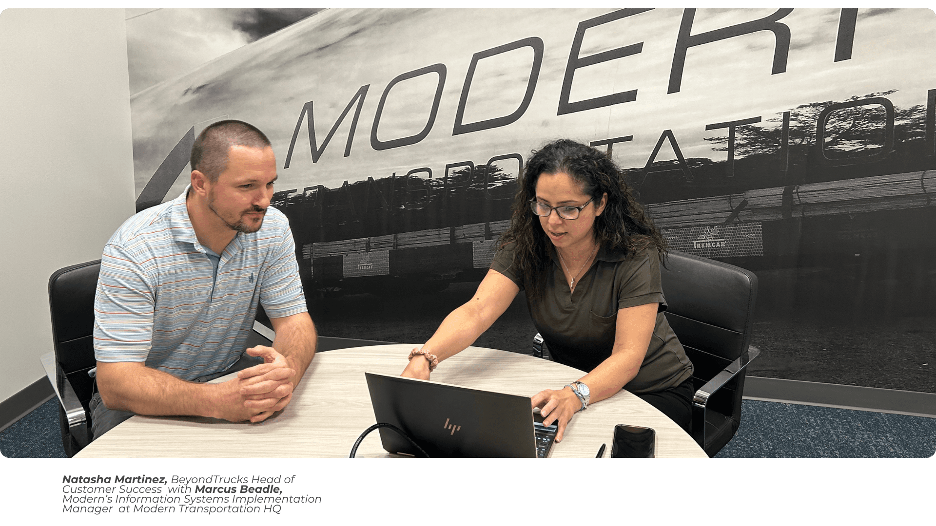 A couple sits at a round office table. The woman is wearing a black shirt and typing on her laptop. Next to her, the man in a light-colored dress shirt, with a beard and short hair, is watching the woman. Behind her, on the wall of the room, you can read in large letters the name of the company with the stylized logo "Modern Transportation."
