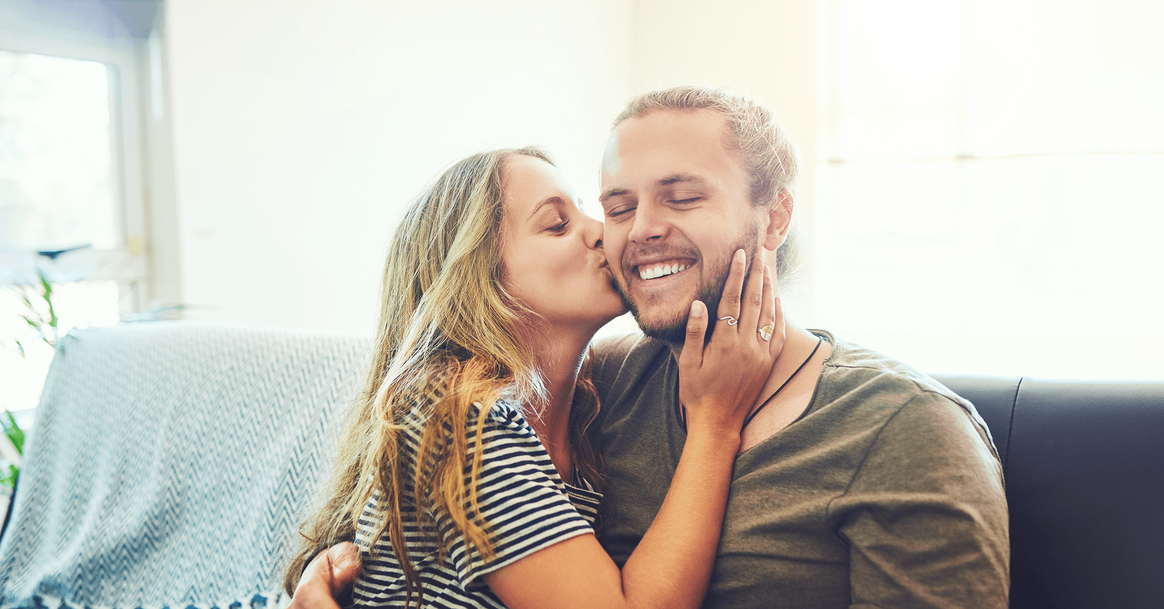 Ett par sitter i soffan. Kvinnan kysser mannen på kinden medan hon håller sin ena hand på hans ansikte. Mannen ler brett med stängda ögon medan han håller om henne.