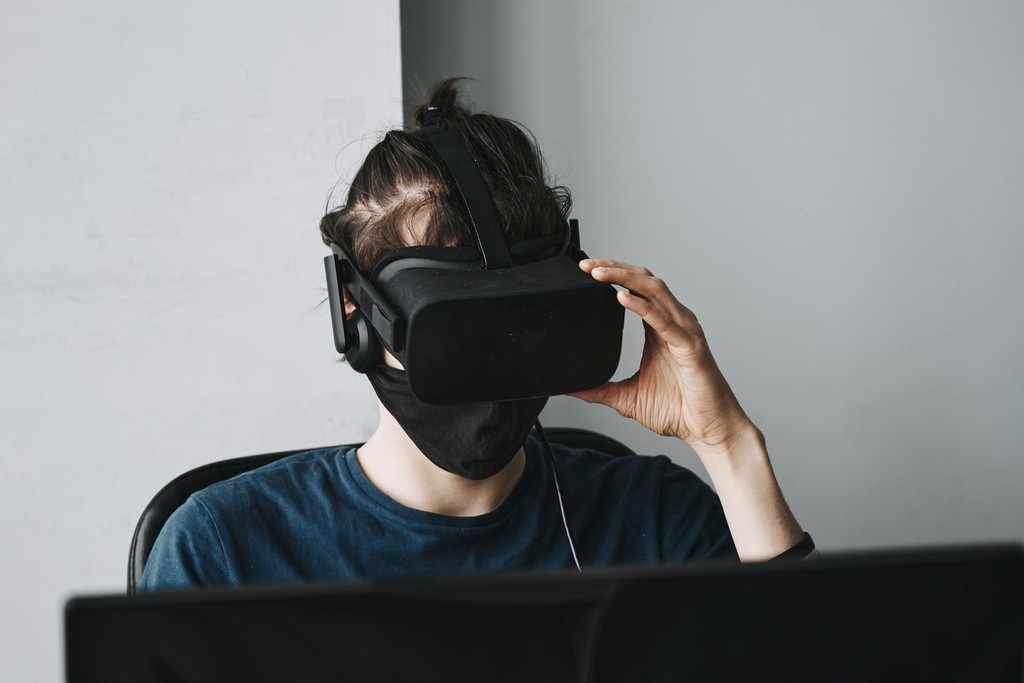 A person wearing a virtual reality headset and a face mask sits at a desk, immersed in a VR experience, highlighting the blend of advanced technology and contemporary health practices.