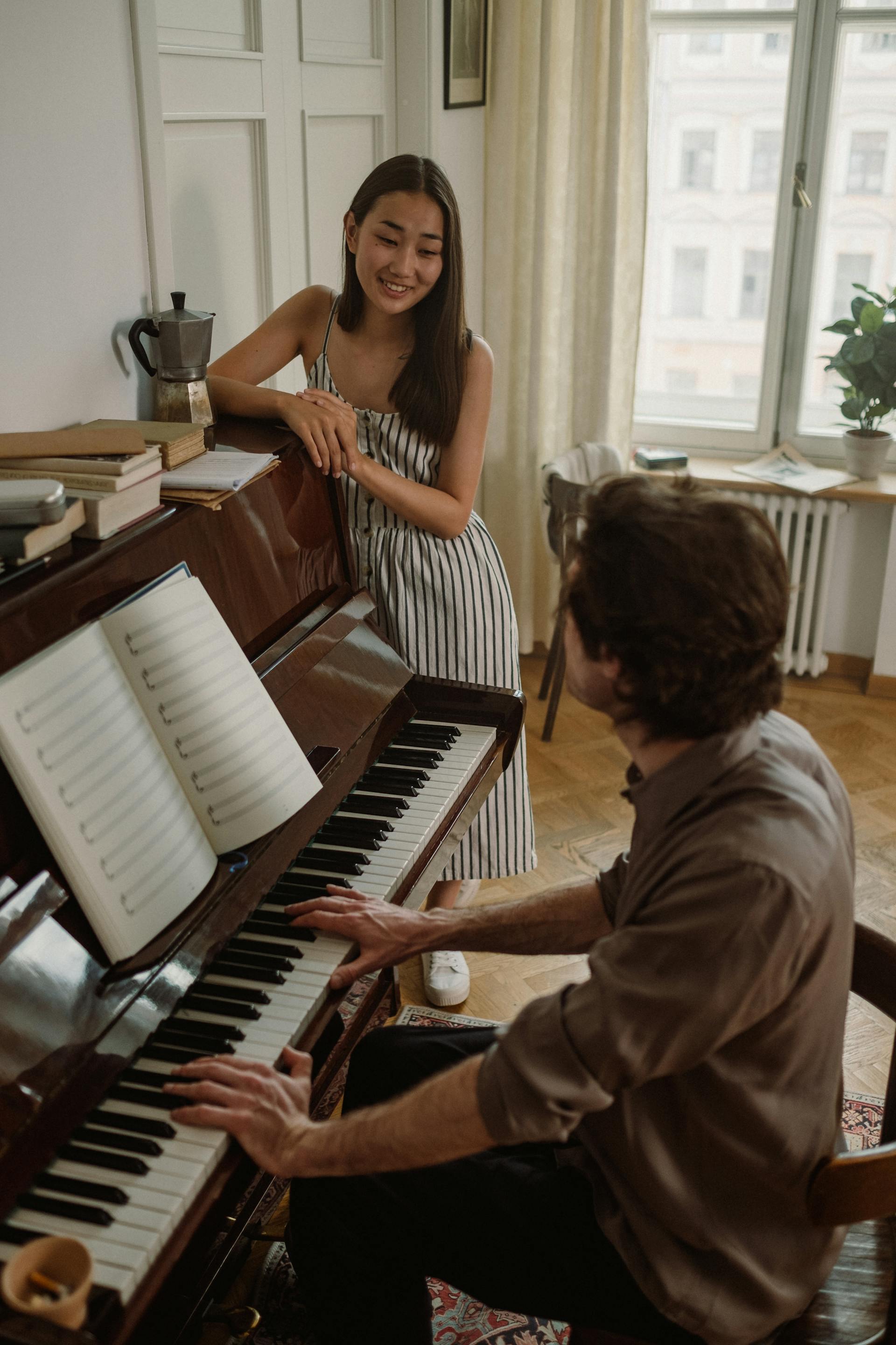 A woman leans on an upright piano, smiling at a man who is seated and playing from a book of sheet music.