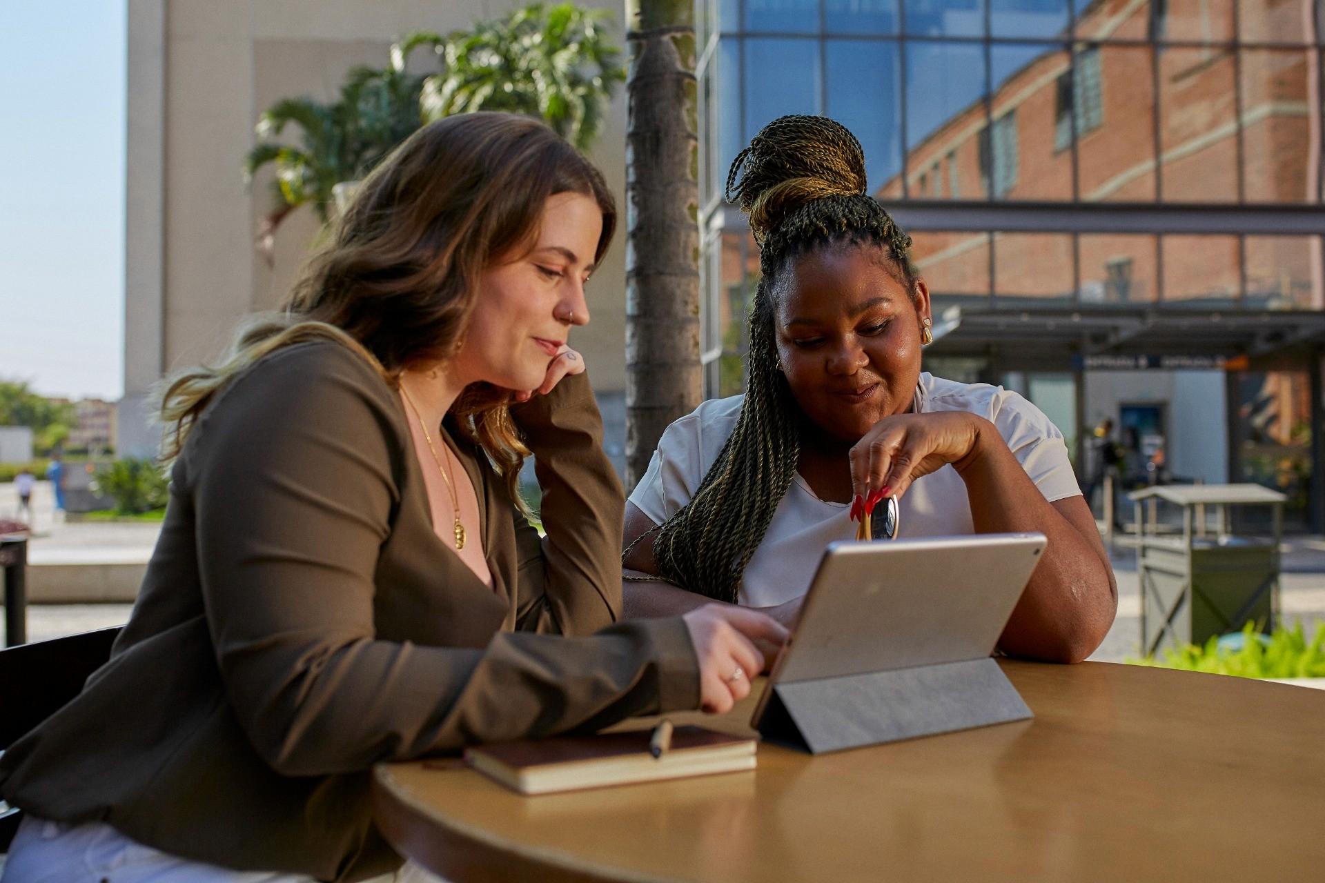 Esta imagem mostra duas mulheres sentadas em uma mesa ao ar livre, trabalhando ou discutindo algo em um tablet. A mulher à esquerda tem cabelo castanho claro, veste uma blusa de manga longa verde-oliva e está observando a tela do dispositivo com interesse. Ela tem um caderno fechado à sua frente. A mulher à direita, com tranças, veste uma blusa clara e está sorrindo enquanto segura um objeto próximo à tela do tablet, possivelmente uma caneta stylus. Atrás delas, há uma estrutura moderna de vidro e um ambiente arborizado, sugerindo que estão em um espaço público ou corporativo.