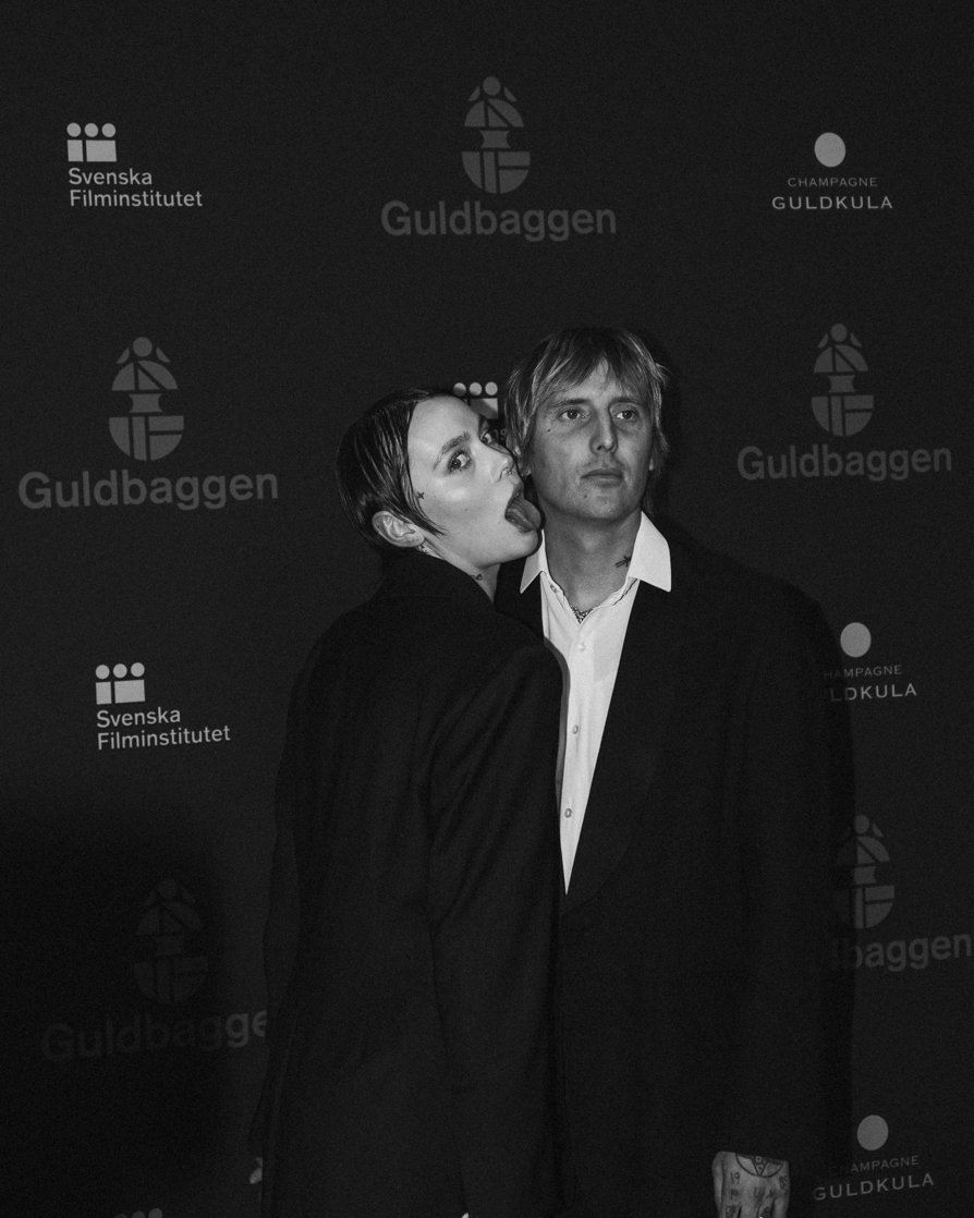 Black and white photo of two people at an event, standing in front of a backdrop with logos and text including 'Guldbaggen' and 'Svenska Filminstitutet.' One person has dark hair and is playfully sticking out their tongue, while the other has light h