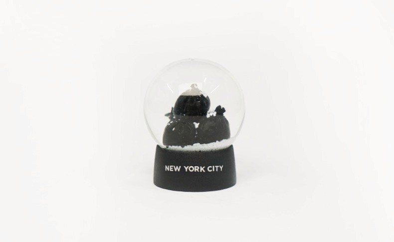 A clear glass snow globe sits on a black base. The base has the words “NEW YORK CITY” written in white. The globe contains a small black figure and a small amount of white material at the bottom. The background is plain white.