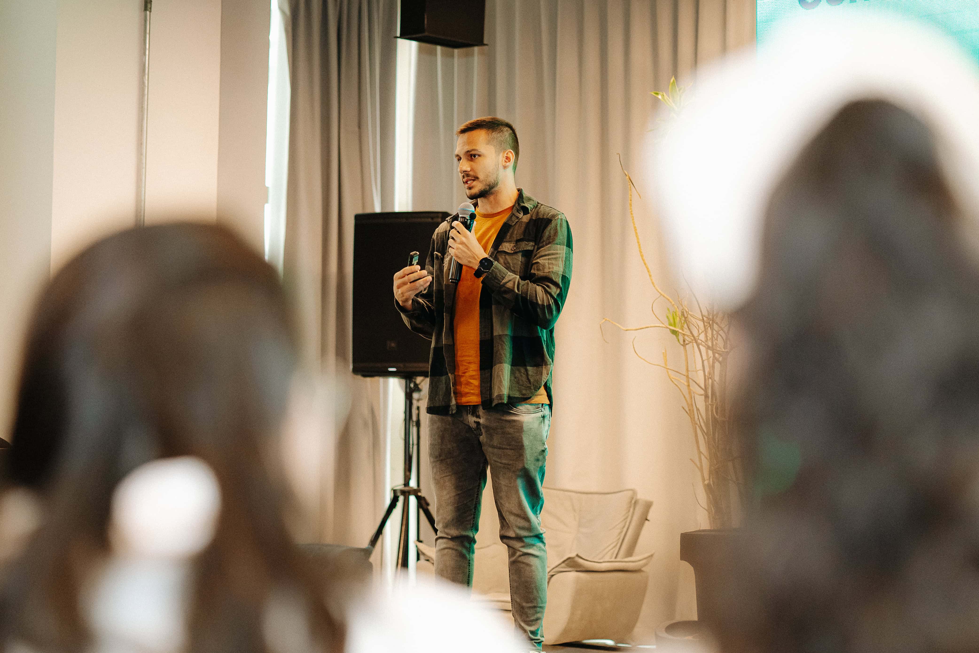 Uma pessoa está em pé no palco falando ao microfone, vestindo uma camiseta preta, com uma planta em primeiro plano.