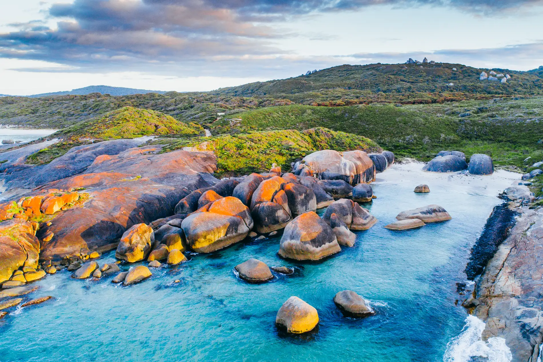 Aerial of orange-lichened granite boulders along the water's edge below green coastal hills