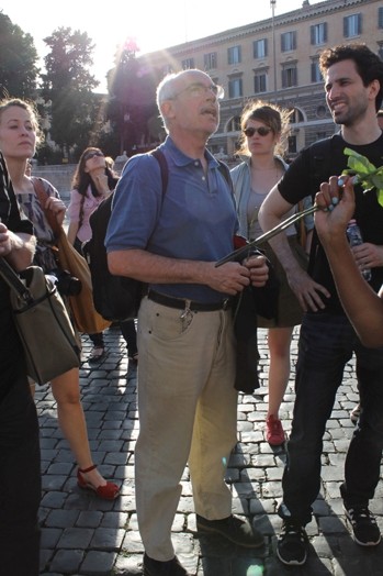 A photo of a man giving a tour and standing among other people in a roman paved old square.