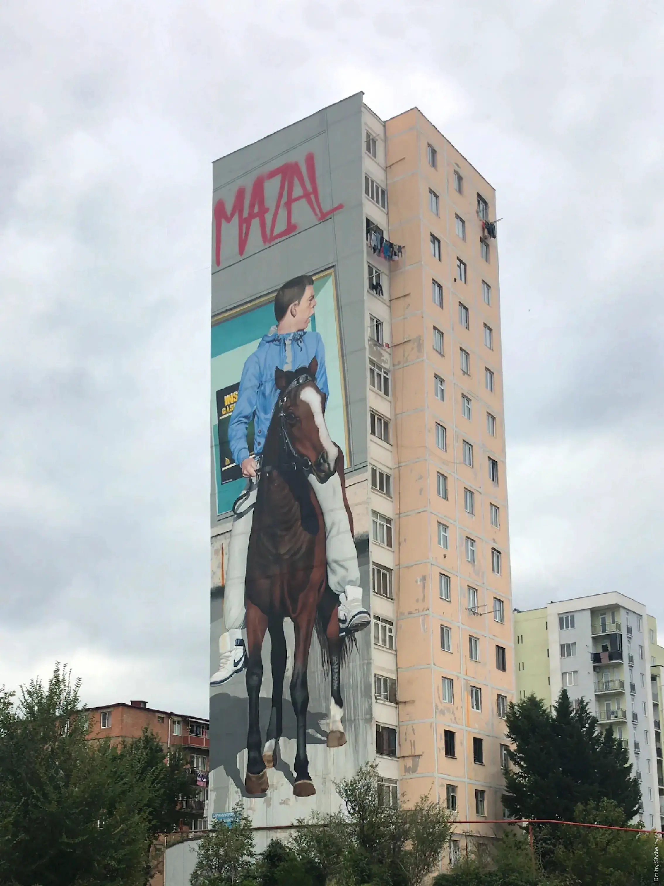 A photorealistic mural on the narrow side of a tall Soviet-era apartment building, depicting a young man in a blue shirt sitting atop a brown and white horse.