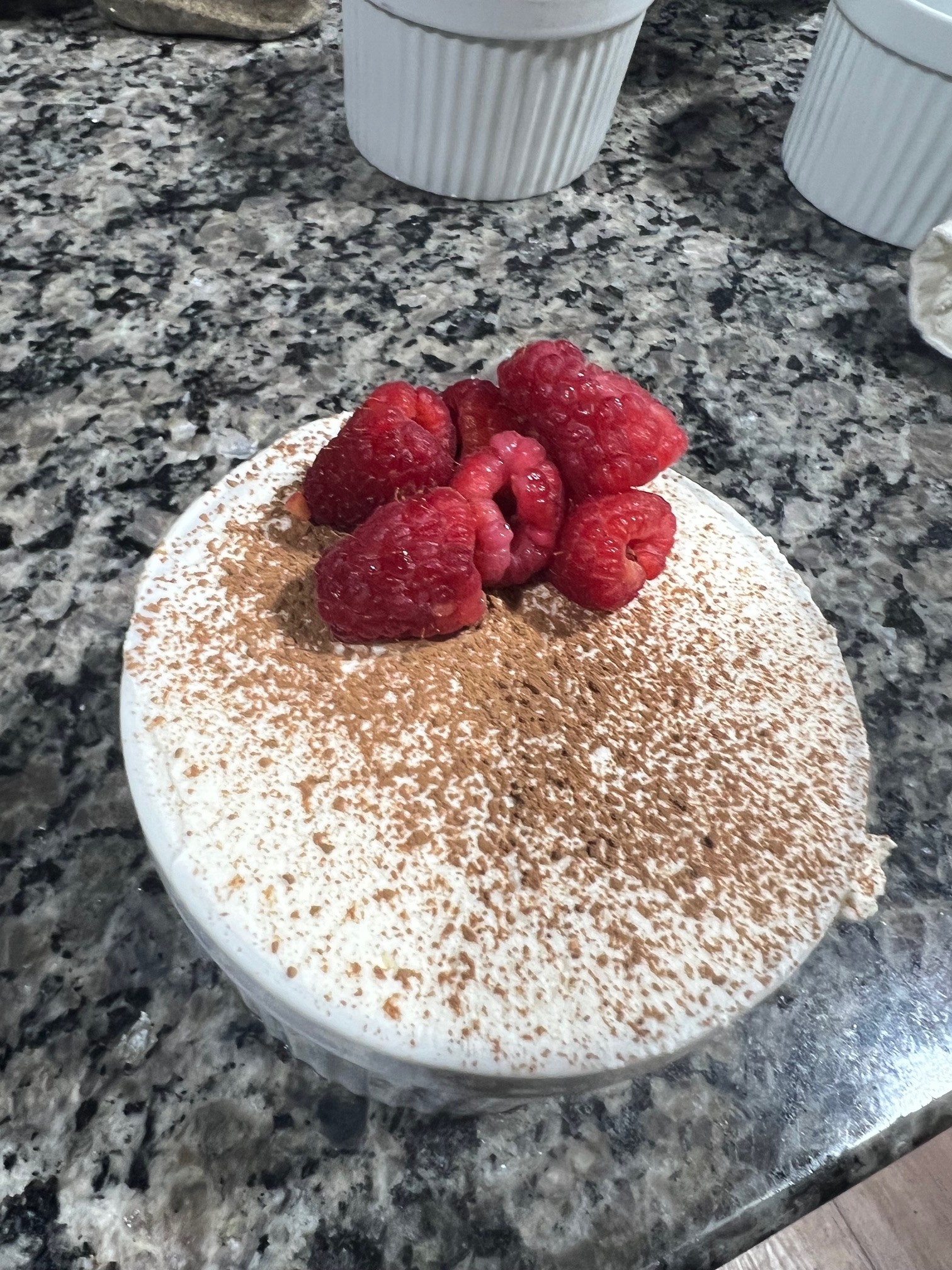 Dessert in white bowl on granite surface, raspberries on top, sprinkled with brown powder.