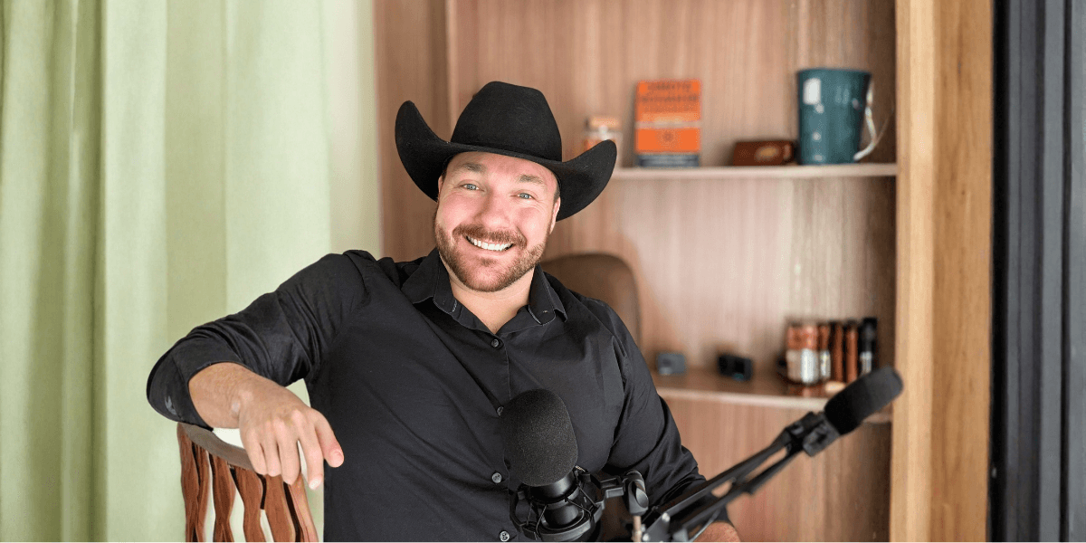 a man wearing a cowboy hat, smiling while sitting beside a microphone in a podcast setup.