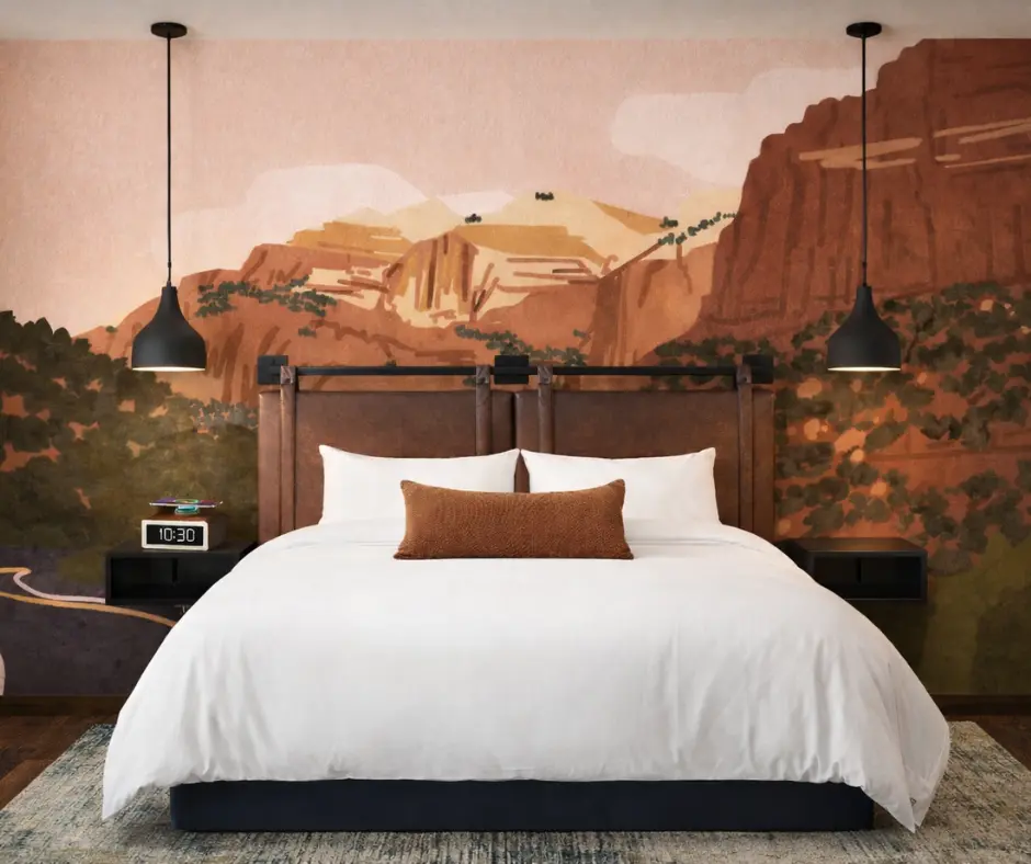 A modern bedroom featuring a white bed, brown accents, and a mural of mountains in warm hues.