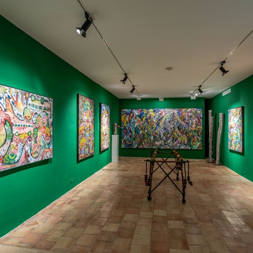 Art gallery with green walls displaying colorful abstract paintings and sculptures. Spotlights illuminate the artworks.
